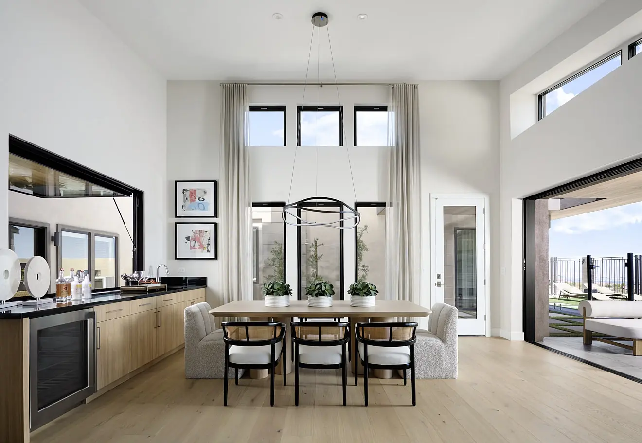 250911_Carlisle_Ridge_P2_003 Modern dining room in Plan 2 at Carlisle Ridge by Tri Pointe Homes, featuring light wood floors, a large table for six, potted plant centerpiece, high ceilings, tall windows with light drapes, minimalist black chandelier, and glass doors to the outdoors.