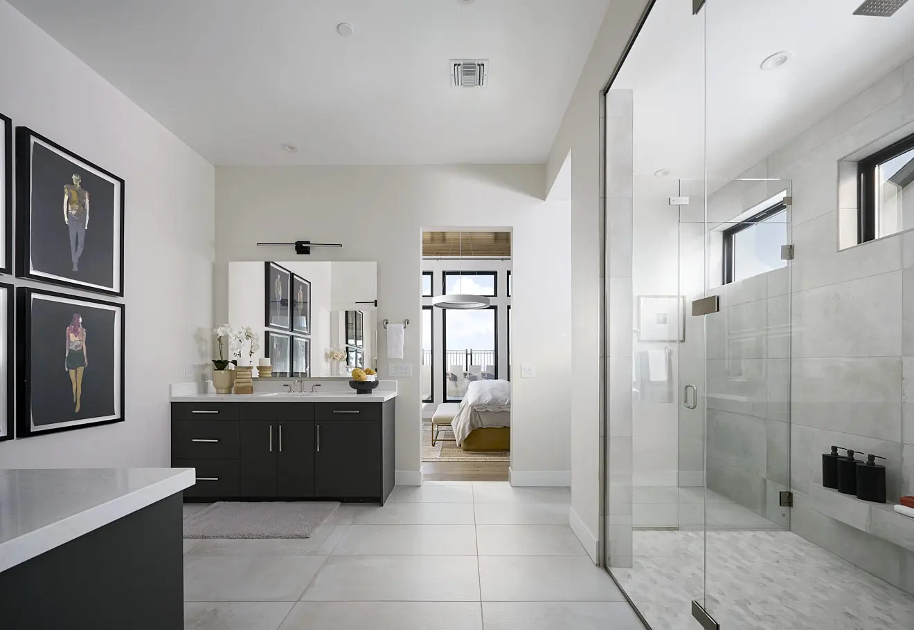 250911_Carlisle_Ridge_P2_006 Modern bathroom in Plan 2 at Carlisle Ridge by Tri Pointe Homes features a large glass shower, double-sink dark vanity, wall art, white walls, and a view into the bedroom through an open doorway. Natural light streams in from multiple windows.