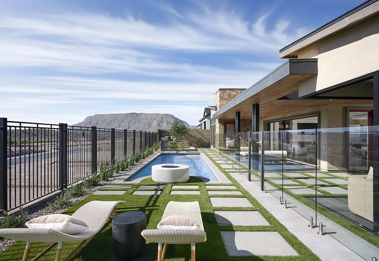 250911_Carlisle_Ridge_P2_007 Modern backyard at Plan 2 at Carlisle Ridge by Tri Pointe Homes with lounge chairs, a small pool, fire pit, and glass fence. Green artificial turf with stepping stones lines the patio, set against mountain views and wispy blue skies.