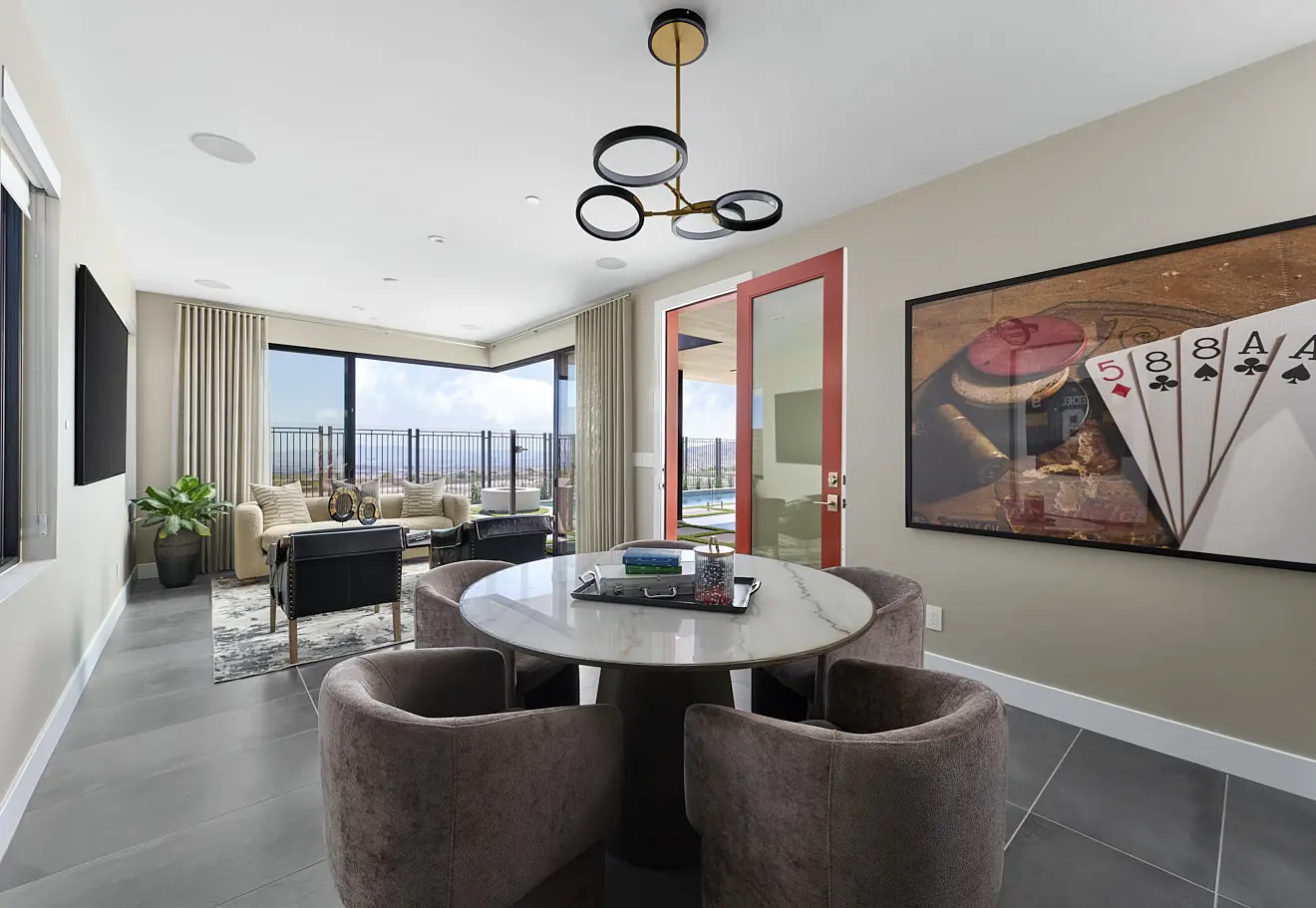 250911_Carlisle_Ridge_P2_011 Modern room in Plan 2 at Carlisle Ridge by Tri Pointe Homes with a round marble table and velvet chairs, a large window leading to a balcony, playing card wall art, plus a lounge area with sofas and plants in the background.