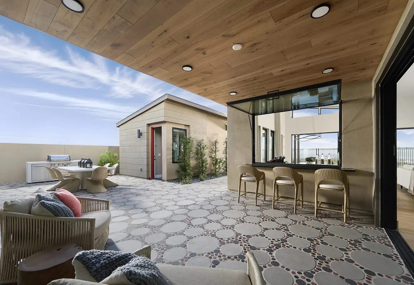 A modern outdoor patio at Plan 2 at Carlisle Ridge by Tri Pointe Homes features patterned tile flooring, wicker furniture, a bar area with stools, a barbecue grill, and potted plants under a wooden-paneled ceiling on a clear, sunny day.