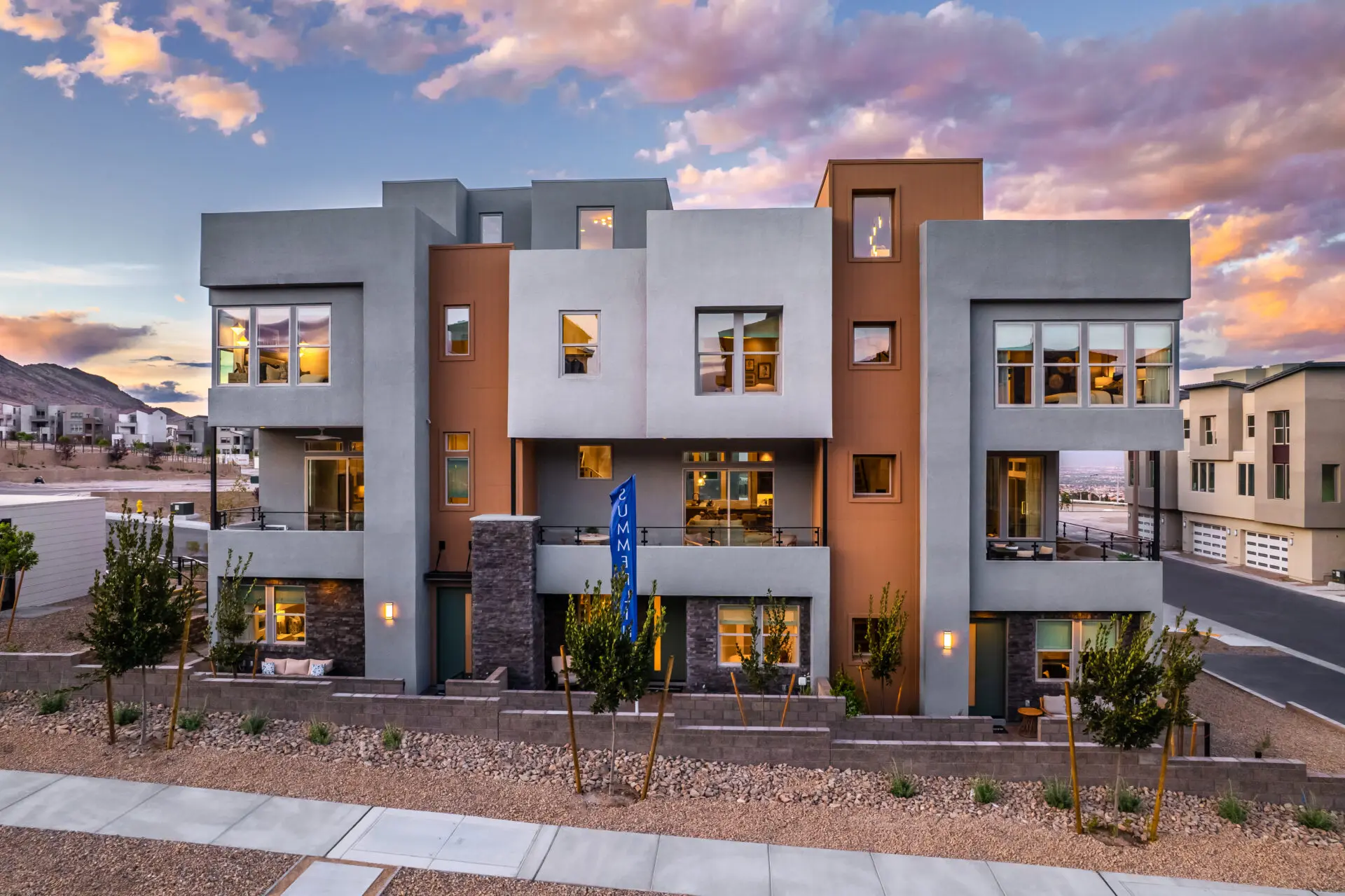 Toll Brothers Raven Crest Modern three-story apartment building with geometric architecture, large windows, and a mix of gray and brown exterior panels, set against a colorful sunset sky and landscaped with small trees and rocks.