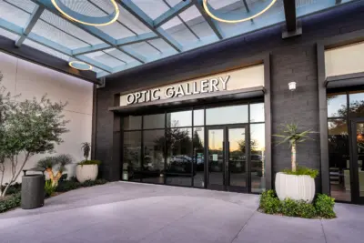 Entrance to a modern building with large glass doors and a sign reading Optic Gallery above. There are potted plants on either side of the doorway and a metal trash can to the left.