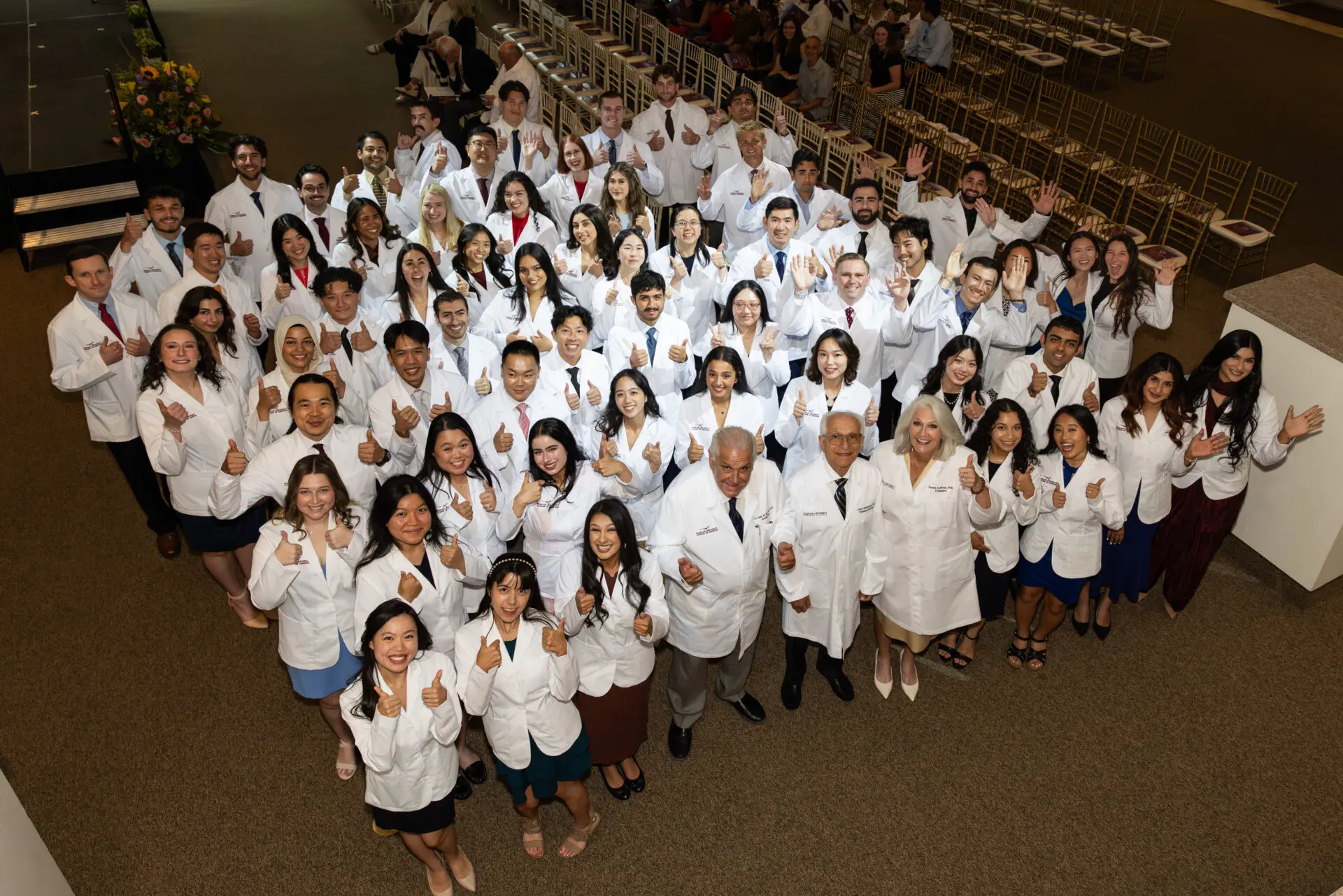 A large group of people in white coats smile and give thumbs up while posing for a group photo indoors, suggesting a celebratory or professional event such as a medical or scientific ceremony.