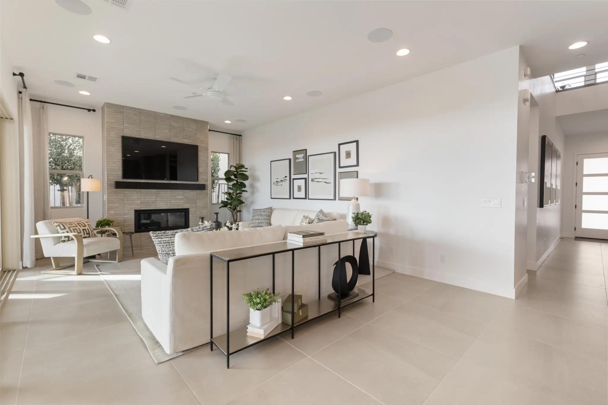 Primrose-Park-Sage-10-2048×1365 A modern, bright living room in the Sage II Floorplan at Primrose Park by Richmond American Homes features a white sofa, black console table, wall-mounted TV above a fireplace, large windows, neutral decor, plants, and framed art. Natural light fills the space.