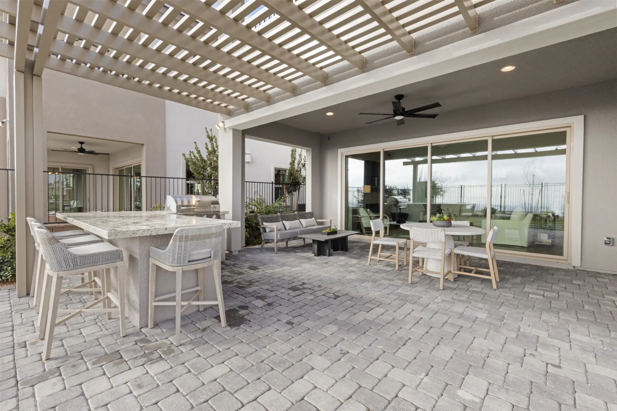 Primrose-Park-Sage-11-2048×1365 Modern outdoor patio with a pergola, bar area with stools, grill, sofa seating, dining table, and paver flooring—just one highlight of the Sage II Floorplan at Primrose Park by Richmond American Homes. Ceiling fans and greenery complete this open-air space.