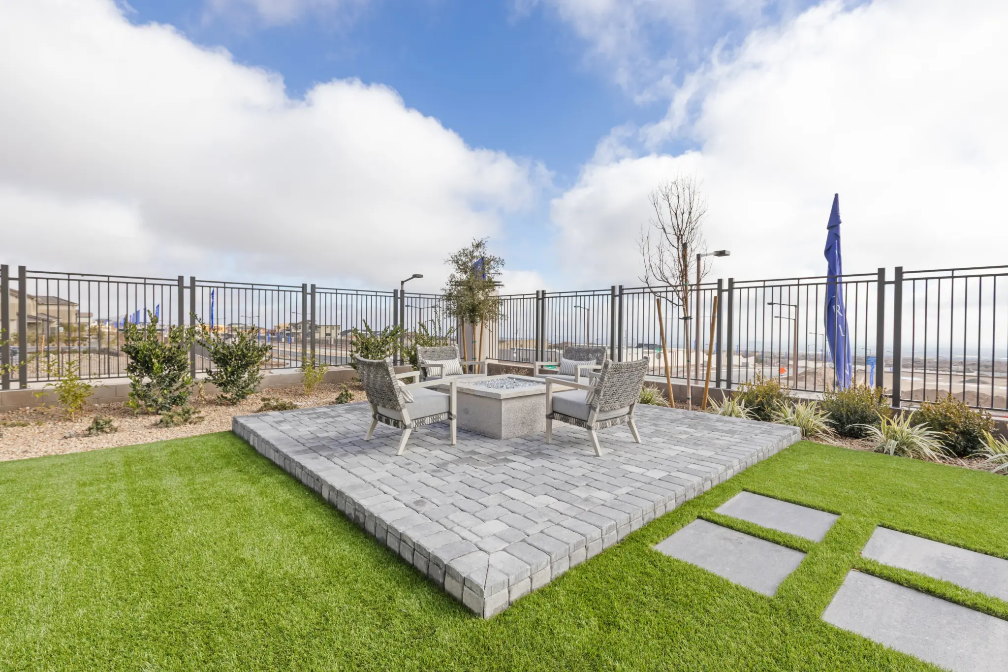 Primrose-Park-Sage-12-2048×1365 A modern backyard patio in the Sage II Floorplan at Primrose Park by Richmond American Homes features four chairs around a square fire pit on a raised stone platform, surrounded by green grass, landscaping, and a metal fence under a partly cloudy sky.