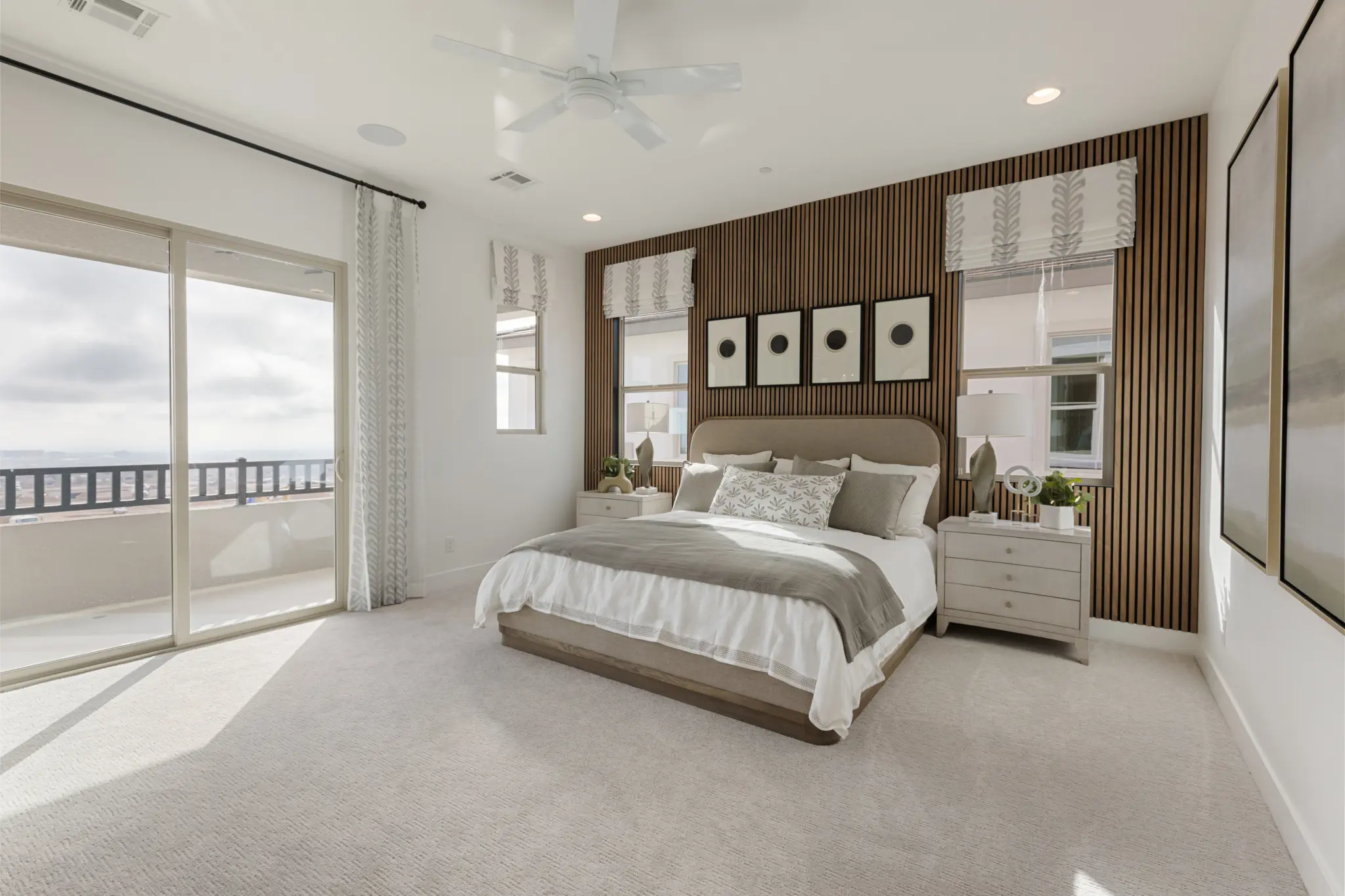 Primrose-Park-Sage-2-2048×1365 A modern, bright bedroom in the Sage II Floorplan at Primrose Park by Richmond American Homes features a large bed, neutral bedding, two nightstands, wall art above the headboard, tall windows, sliding glass doors to a balcony, and light carpeted floors.