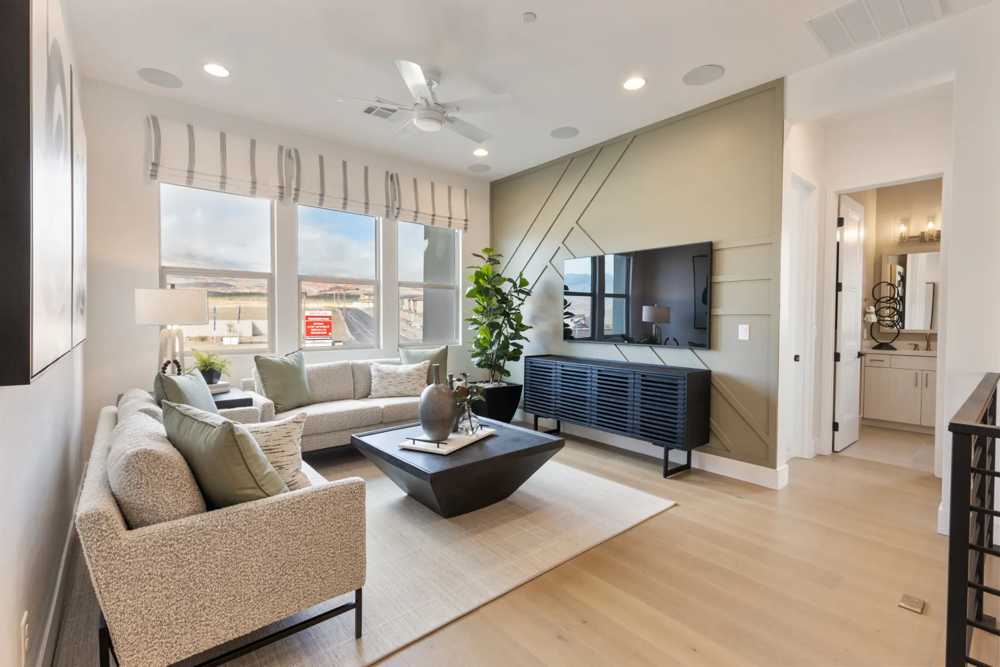 Primrose-Park-Sage-5-2048×1365 A modern living room in the Sage II Floorplan at Primrose Park by Richmond American Homes features light wood floors, a beige rug, two sofas, a black coffee table, a wall-mounted TV, and large windows. A bathroom is visible through an open door.