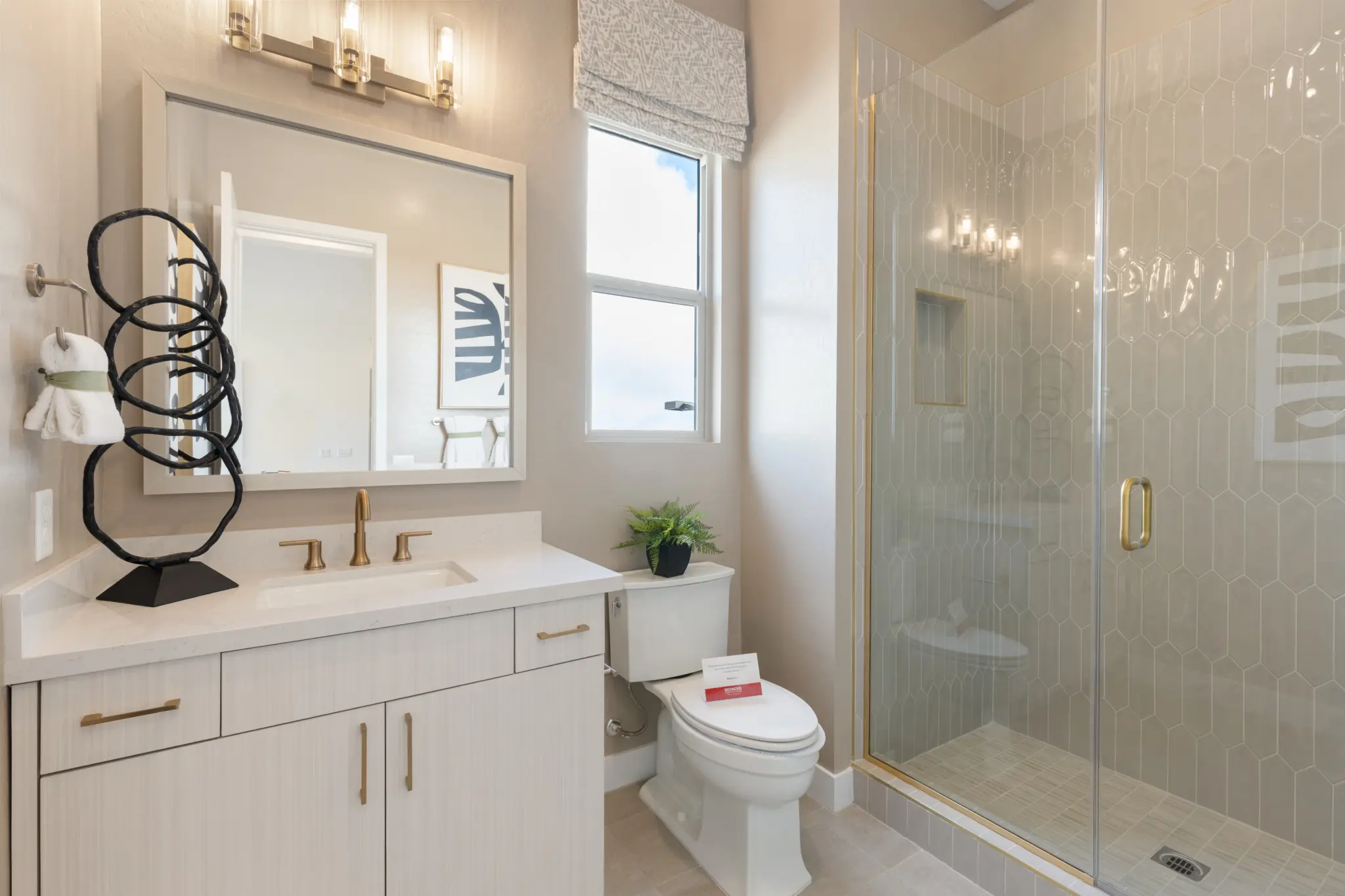 Primrose-Park-Sage-6-2048×1365 Modern bathroom in the Sage II Floorplan at Primrose Park by Richmond American Homes featuring a glass shower, white vanity with gold fixtures, mirror, decorative sculpture, small plant on the toilet tank, and natural light from a window.
