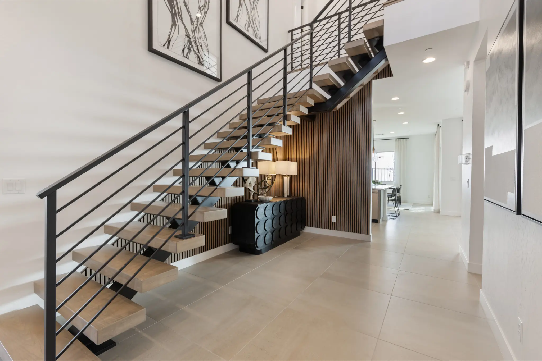 Primrose-Park-Sage-7-2048×1365 Modern interior with a floating wooden staircase and black metal railing, large tile floor, abstract wall art, and a console table with decor—discover the elegance of the Sage II Floorplan at Primrose Park by Richmond American Homes.