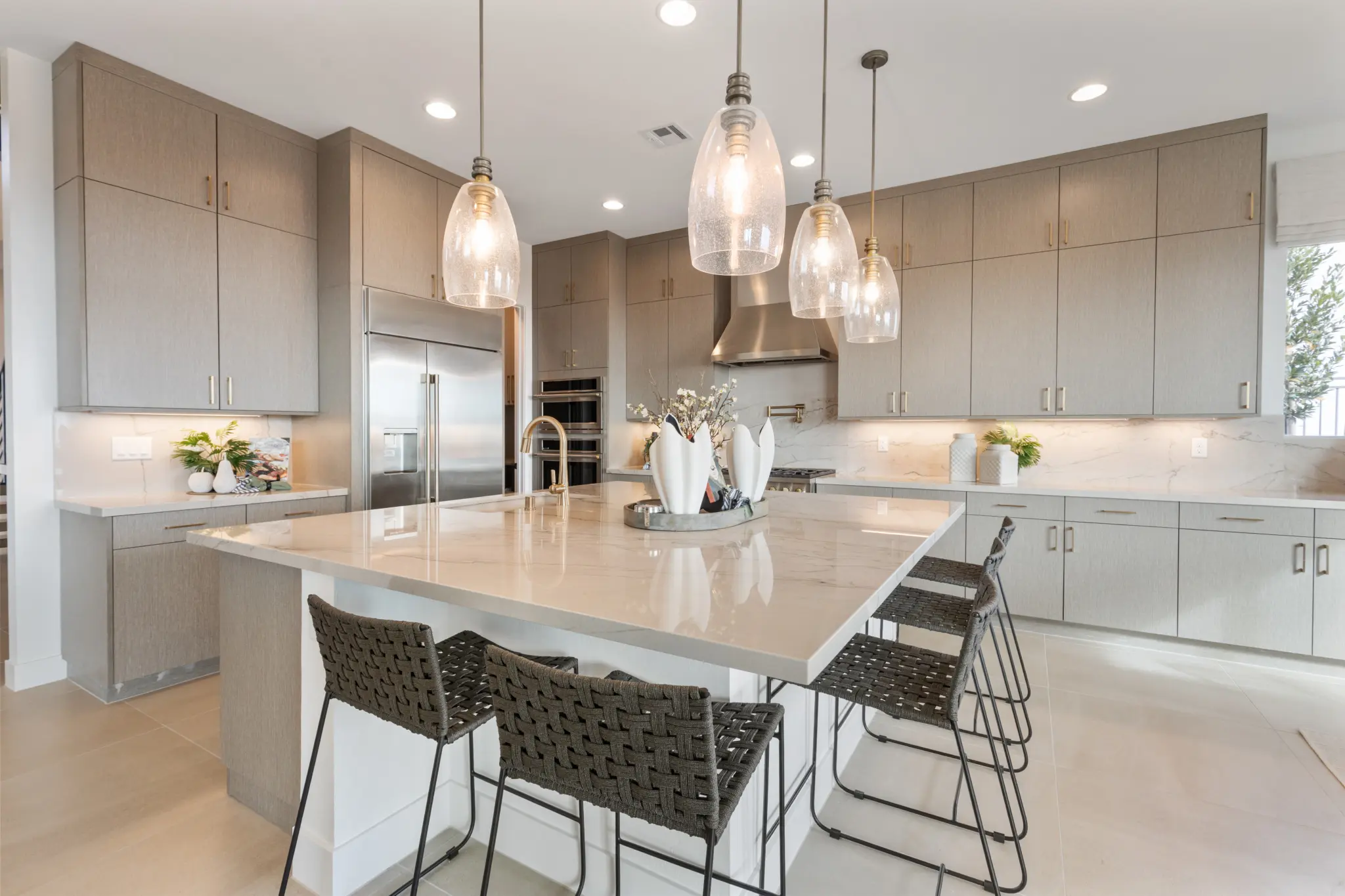 Primrose-Park-Sage-8-2048×1365 Modern kitchen with light wood cabinets, stainless steel appliances, a large marble island with four woven barstools, pendant lights overhead, and decorative items—featured in the Sage II Floorplan at Primrose Park by Richmond American Homes.