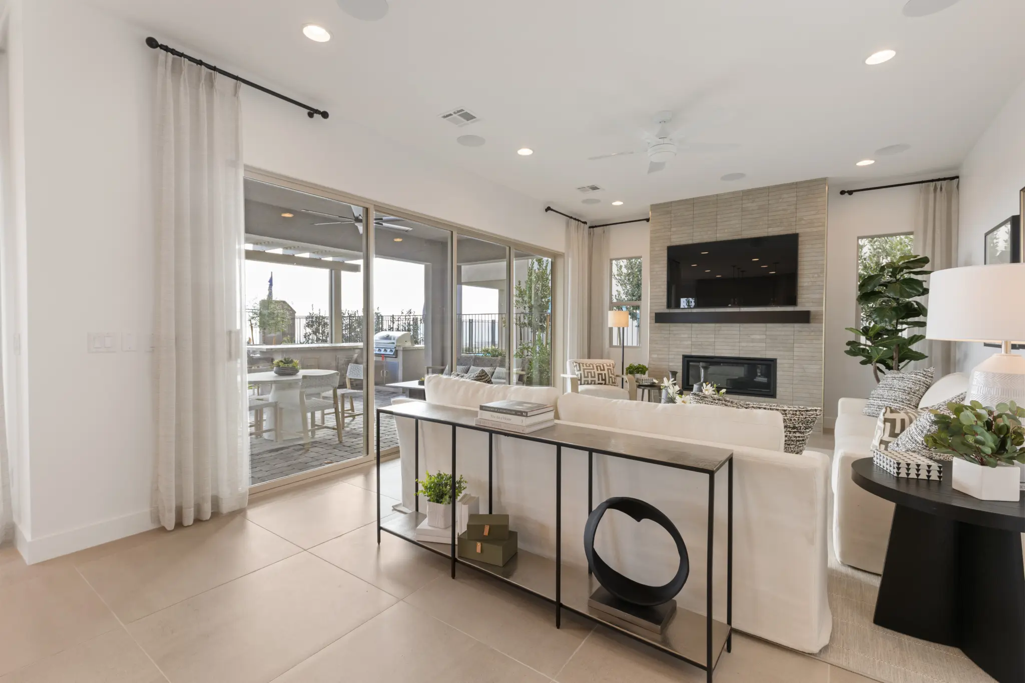 Primrose-Park-Sage-9-2048×1365 A modern living room in the Sage II Floorplan at Primrose Park by Richmond American Homes features a white sofa, wall-mounted TV above a fireplace, light tile floors, large windows with sheer curtains, and a patio view with dining furniture.