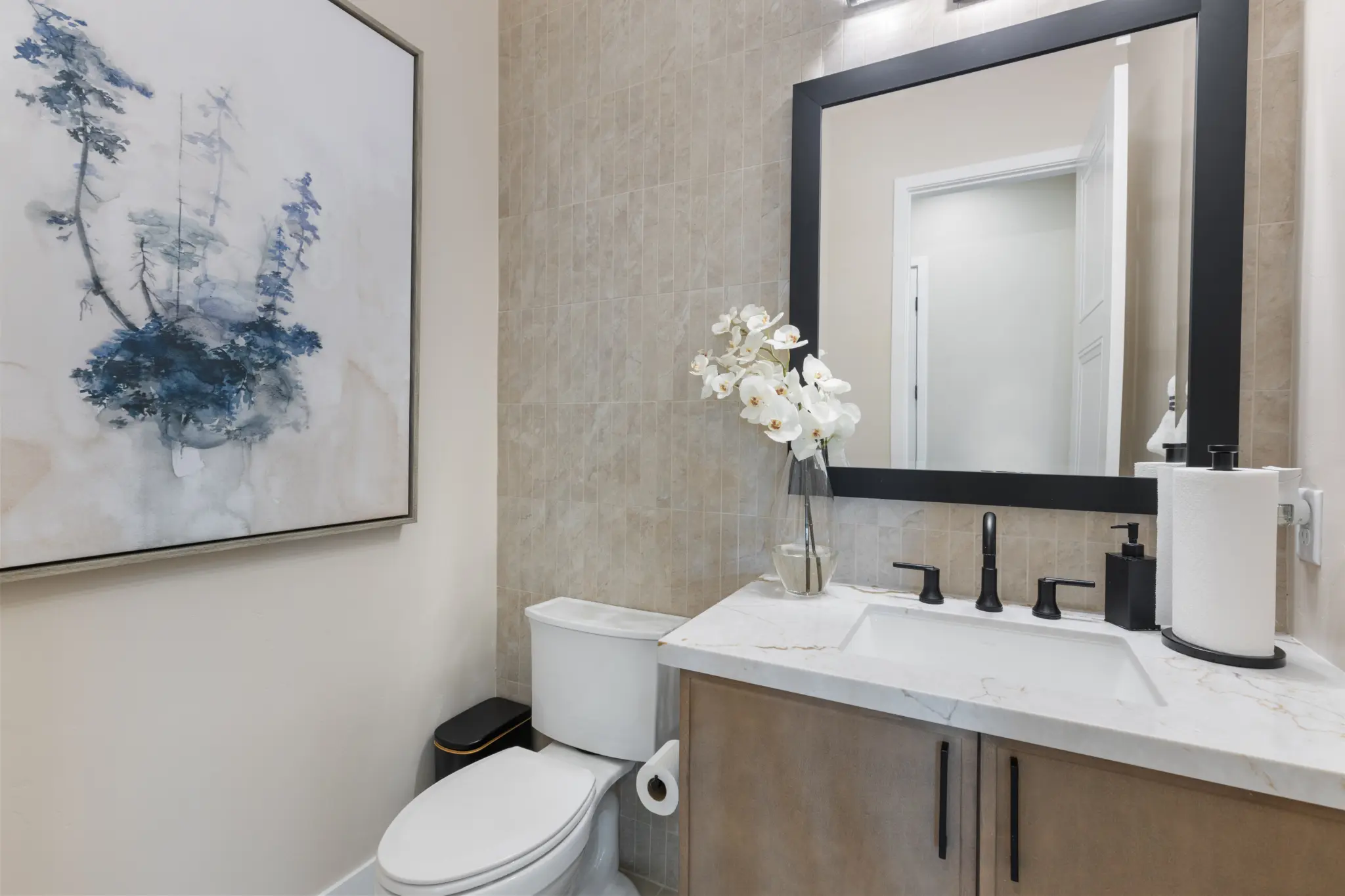Primrose-Park-Teton-1-2048×1365 Modern bathroom in the Teton Floorplan at Primrose Park by Richmond American Homes features a toilet, marble-topped vanity, black fixtures, large mirror, white orchids, a blue abstract painting, neutral tile, and light-colored walls.
