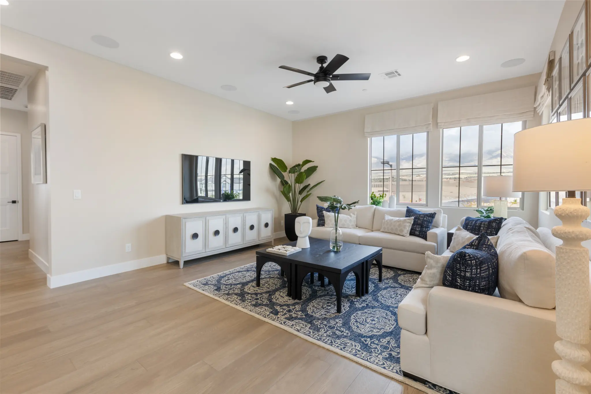 Primrose-Park-Teton-10-2048×1365 Bright, modern living room with light wood floors in the Teton Floorplan at Primrose Park by Richmond American Homes—white walls, a white sofa with blue cushions, blue-patterned rug, black coffee table, wall-mounted TV, large windows, and green potted plants.
