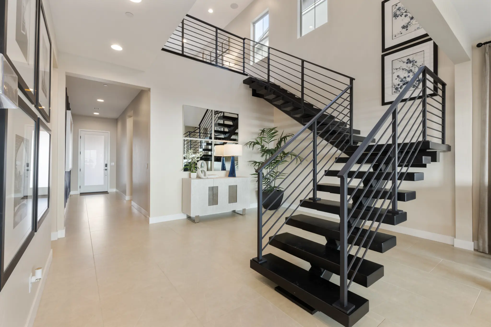 Primrose-Park-Teton-2-2048×1365 Modern entryway with beige tile flooring, open black metal staircase, a hallway to the left, and framed art. The Teton Floorplan at Primrose Park by Richmond American Homes features a console table, indoor plant, and abundant natural light.