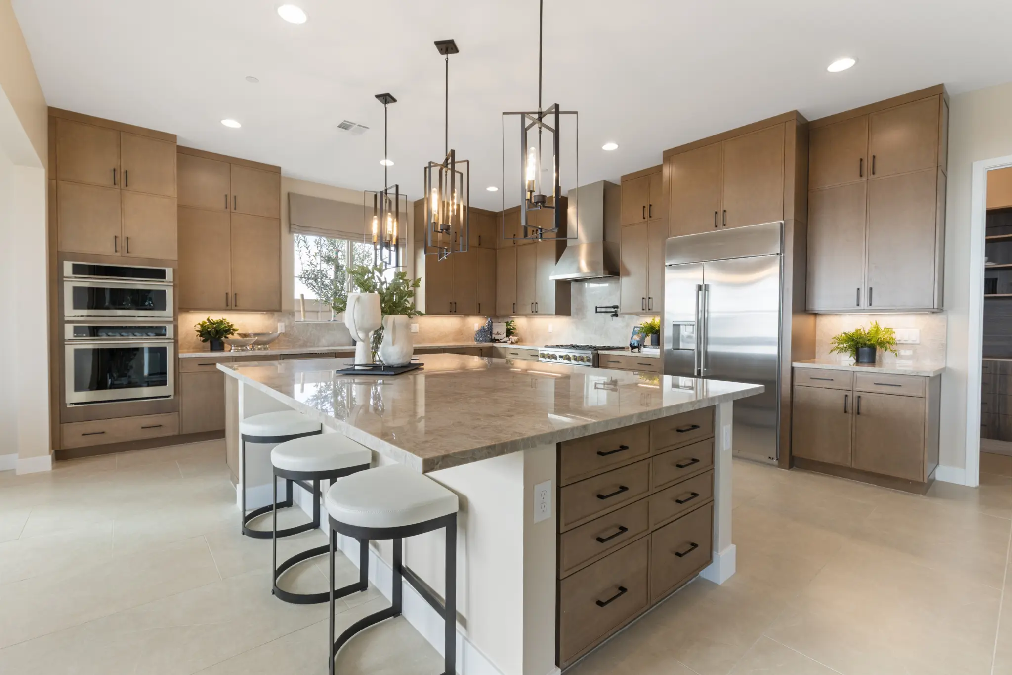 Primrose-Park-Teton-3-2048×1365 Modern kitchen with light wood cabinets, stainless steel appliances, and a large marble island with drawers—features showcased in the Teton Floorplan at Primrose Park by Richmond American Homes. Three white stools, pendant lighting, and decorative plants complete the look.