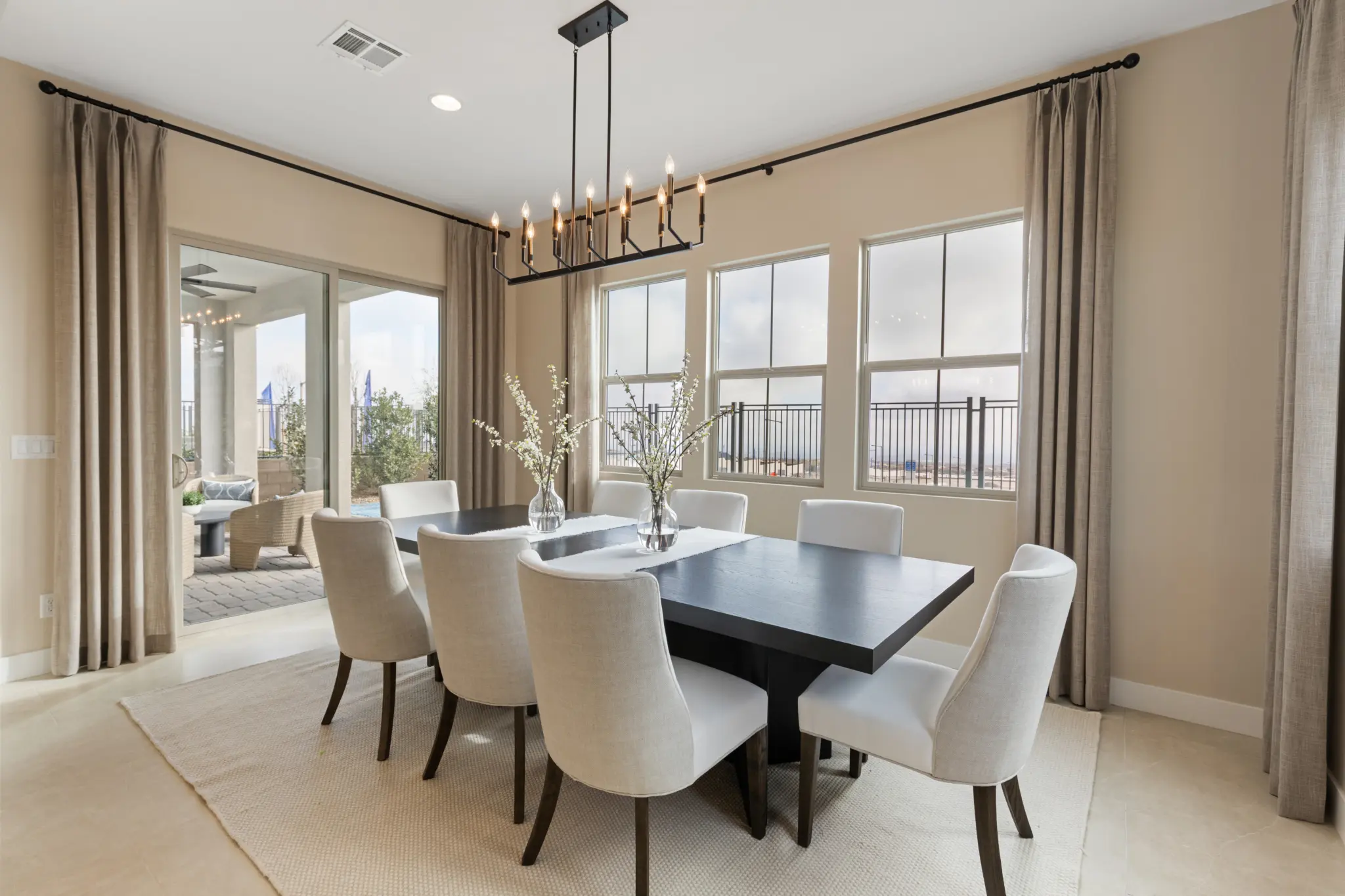 Primrose-Park-Teton-5-2048×1365 A modern dining room in the Teton Floorplan at Primrose Park by Richmond American Homes features a black rectangular table, eight white upholstered chairs, two vases with white flowers, beige walls, large windows, and a black chandelier. Patio visible through open double doors.