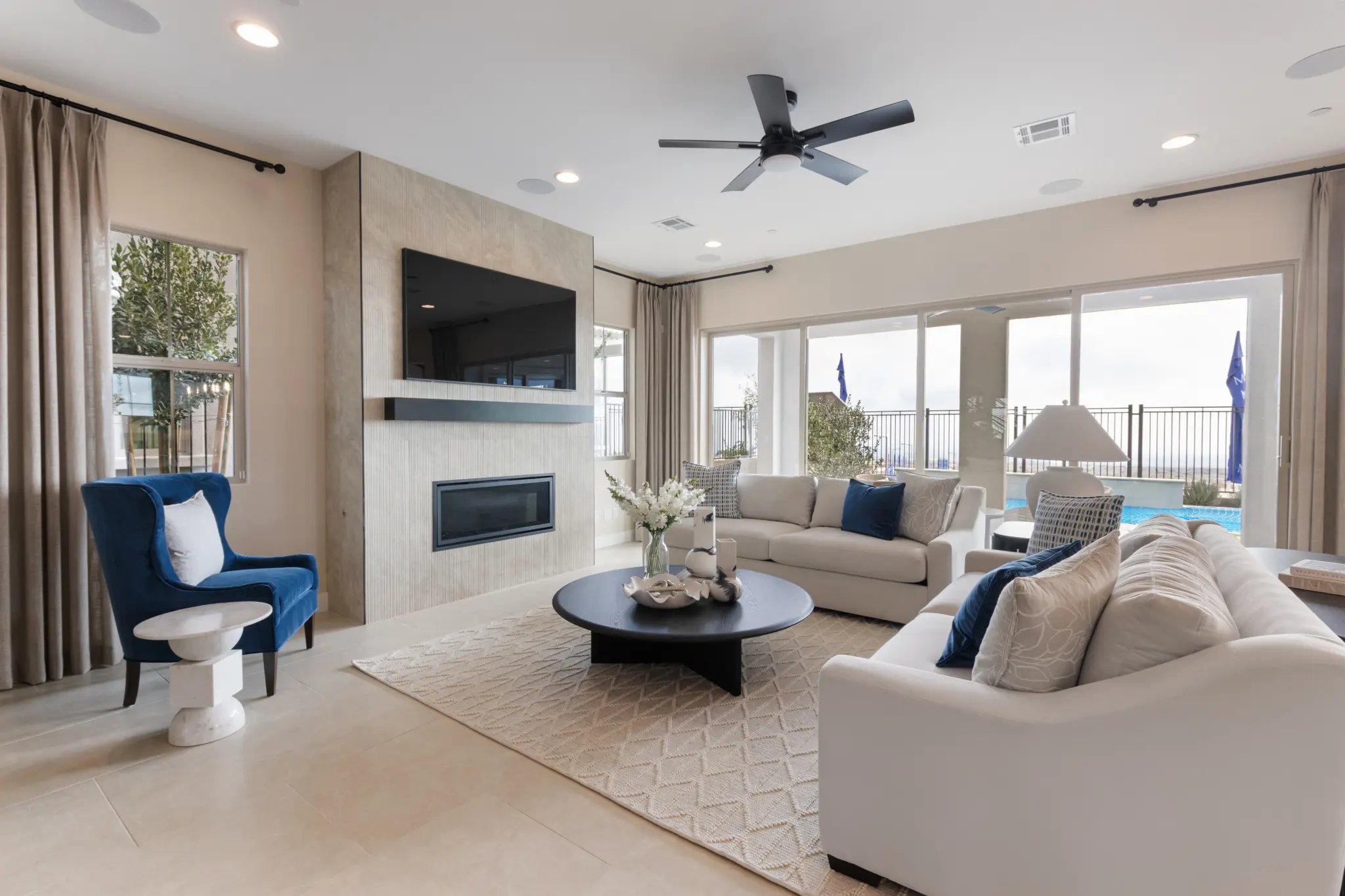 Primrose-Park-Teton-6-2048×1365 Modern living room in the Teton Floorplan at Primrose Park by Richmond American Homes with large windows, a beige sectional sofa, blue armchair, round coffee table, wall-mounted TV above a fireplace, light rug, and views of the pool through glass doors.