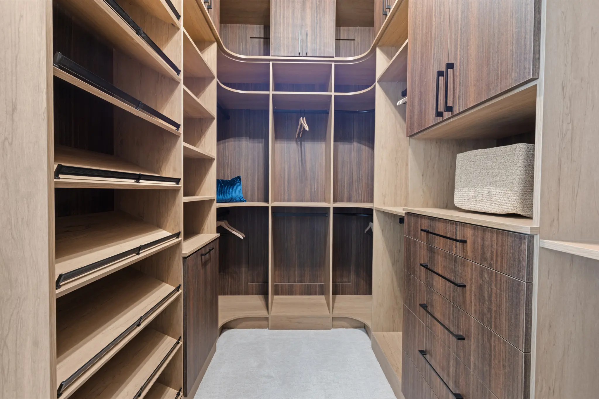Primrose-Park-Teton-8-2048×1365 A spacious walk-in closet in the Teton Floorplan at Primrose Park by Richmond American Homes features wooden shelves, drawers, and cabinets on both sides. There’s a basket, a blue pillow, empty hanging rods, and light grey carpeted flooring.