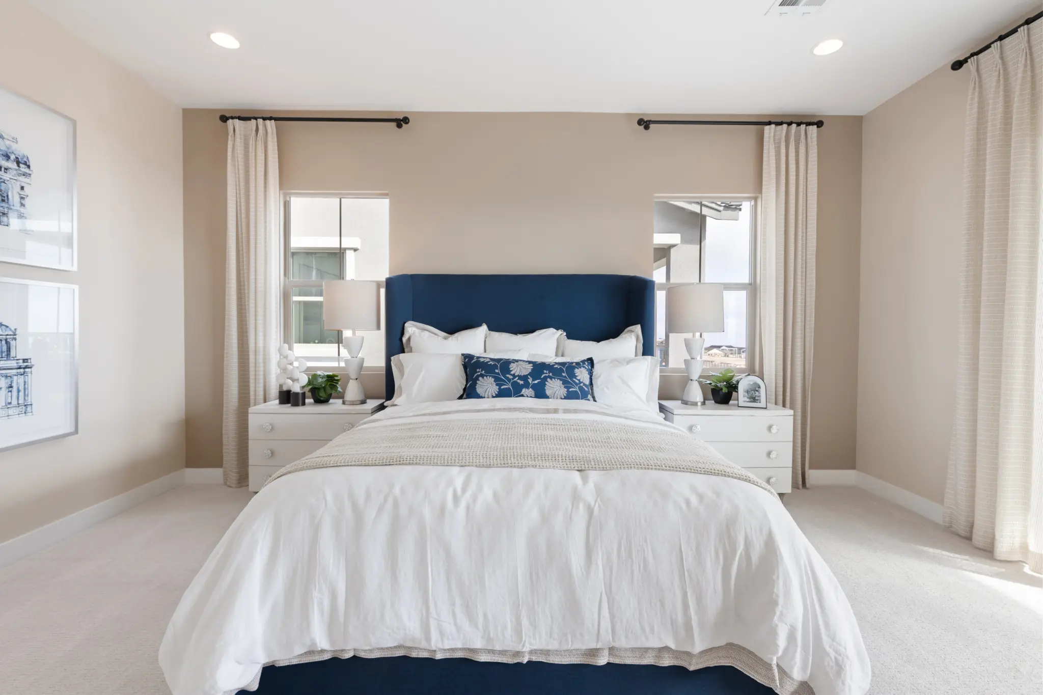 Primrose-Park-Teton-9-2048×1365 A bright, modern bedroom in the Teton Floorplan at Primrose Park by Richmond American Homes features beige walls, a blue upholstered headboard, white bedding, and two nightstands with lamps. Large windows and framed art complete the inviting space.