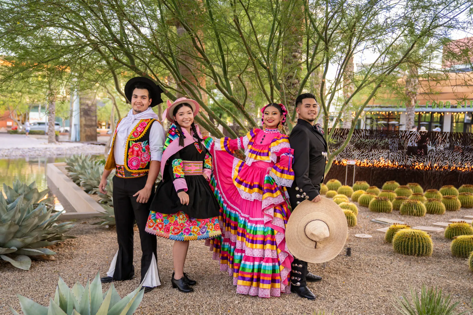 Four people pose outdoors in vibrant traditional Mexican folk dance costumes, surrounded by desert plants and cacti. Two women wear colorful dresses, while two men wear traditional charro suits and hats.