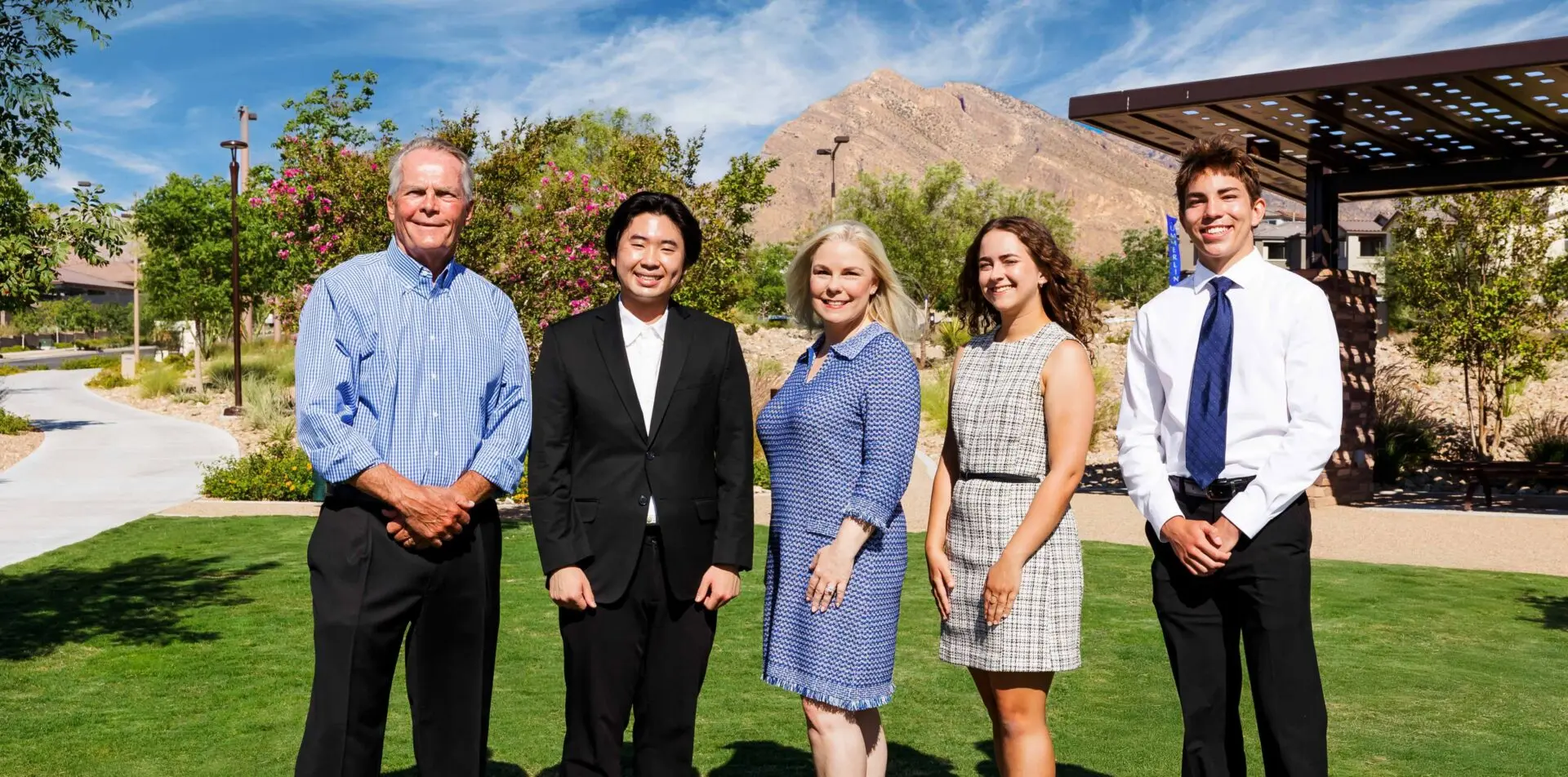 Five people stand outdoors on grass, dressed in business or semi-formal attire, smiling at the camera with a mountain, blue sky, and trees in the background.