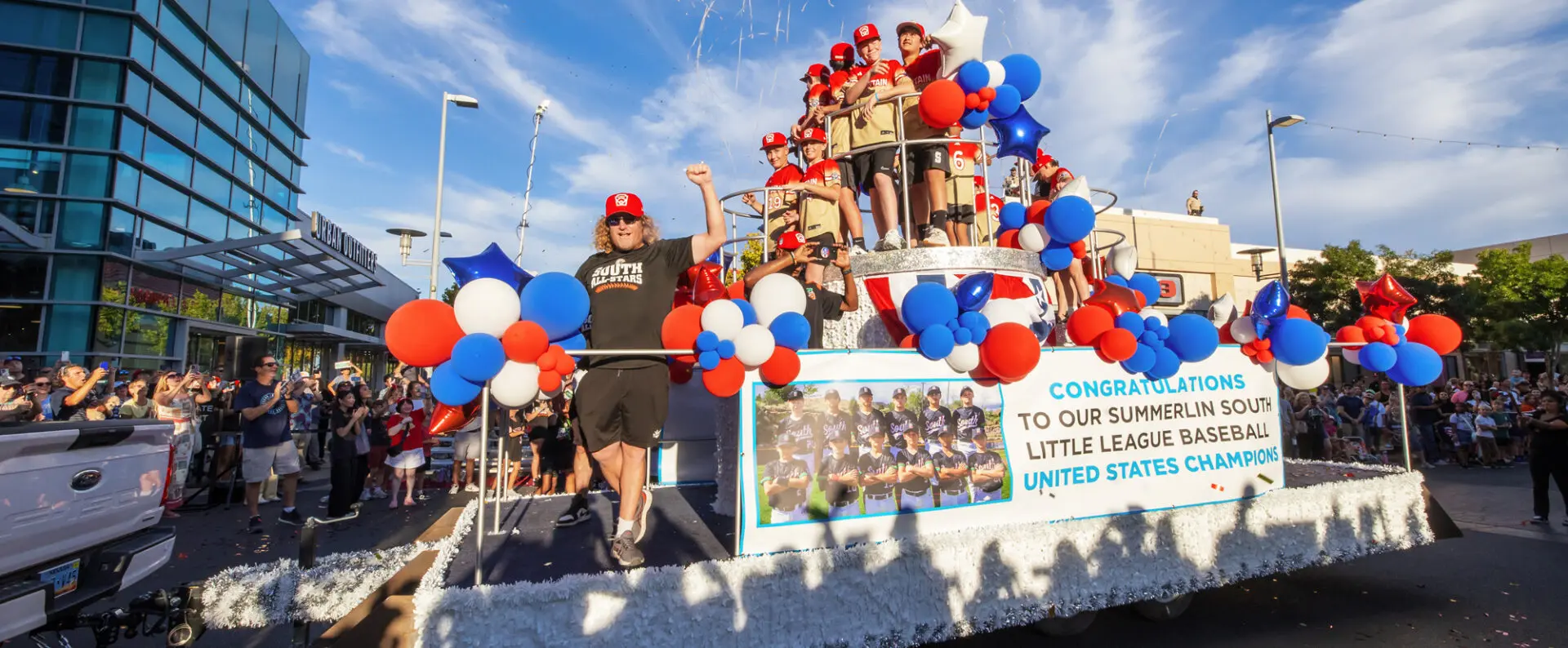 A parade float decorated with red, white, and blue balloons carries a Little League baseball team celebrating their U.S. championship, with cheering fans lining the street and a congratulatory banner on the float.