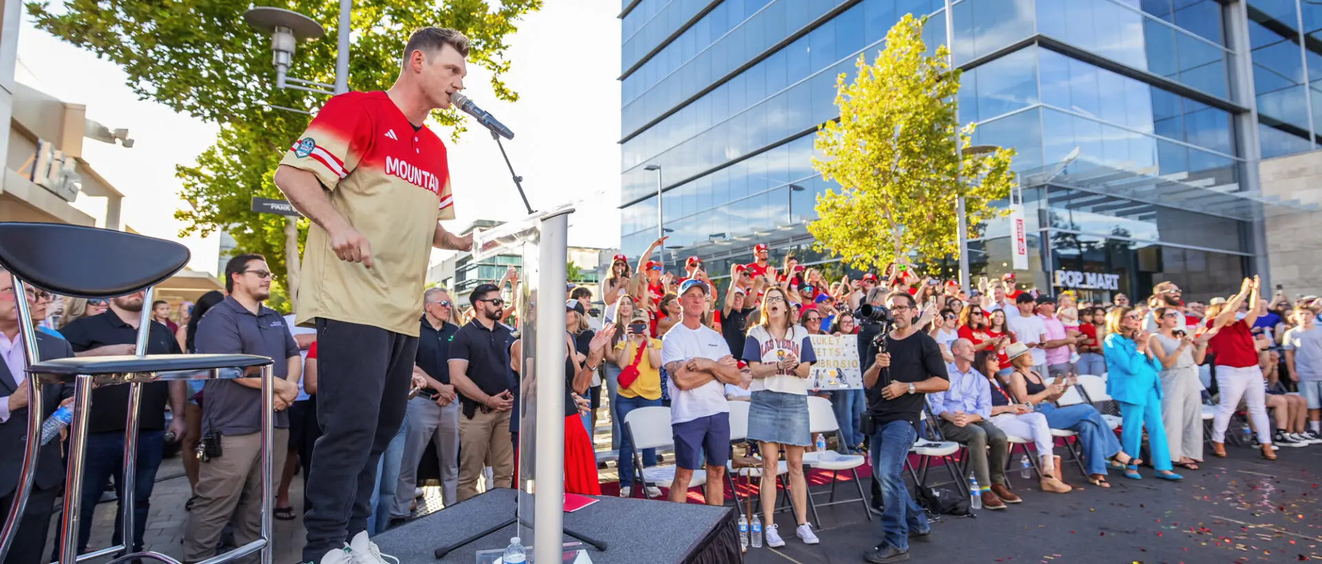 A man in a sports jersey speaks at a podium to a large, enthusiastic outdoor crowd. People stand and sit, some holding cameras or clapping, with a reflective glass building and trees in the background.