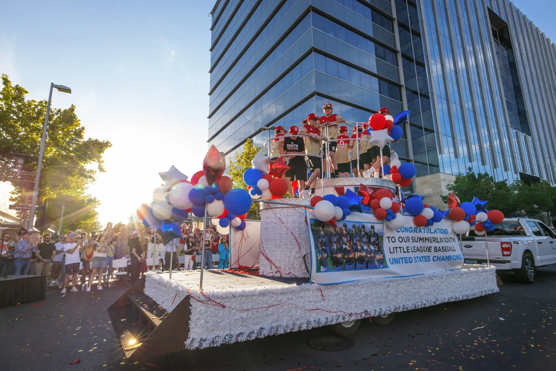 A parade float decorated with red, white, and blue balloons moves down a city street at sunset, carrying people celebrating a little league baseball championship; a crowd watches from the sidewalk.