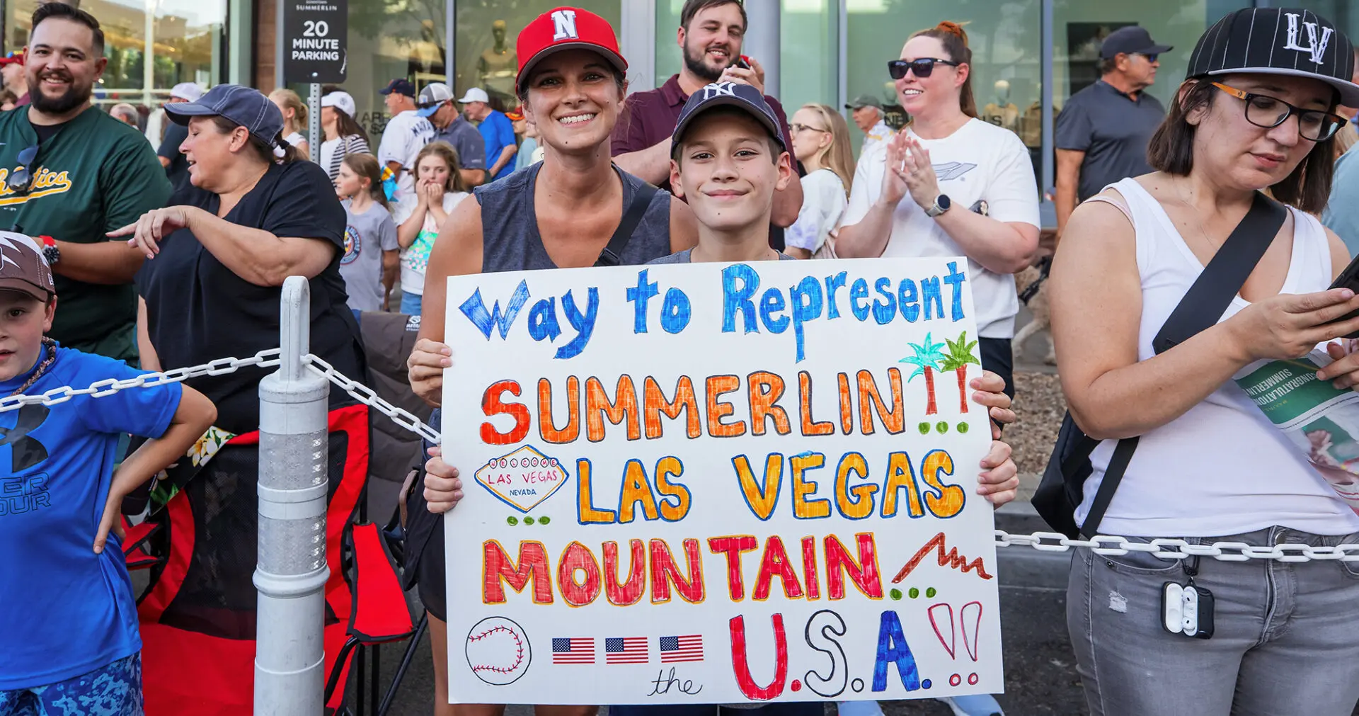 A smiling woman and boy stand in a crowd holding a colorful sign that says “Way to Represent SUMMERLIN!! LAS VEGAS MOUNTAIN, U.S.A.!!” People around them clap and watch, some holding phones.