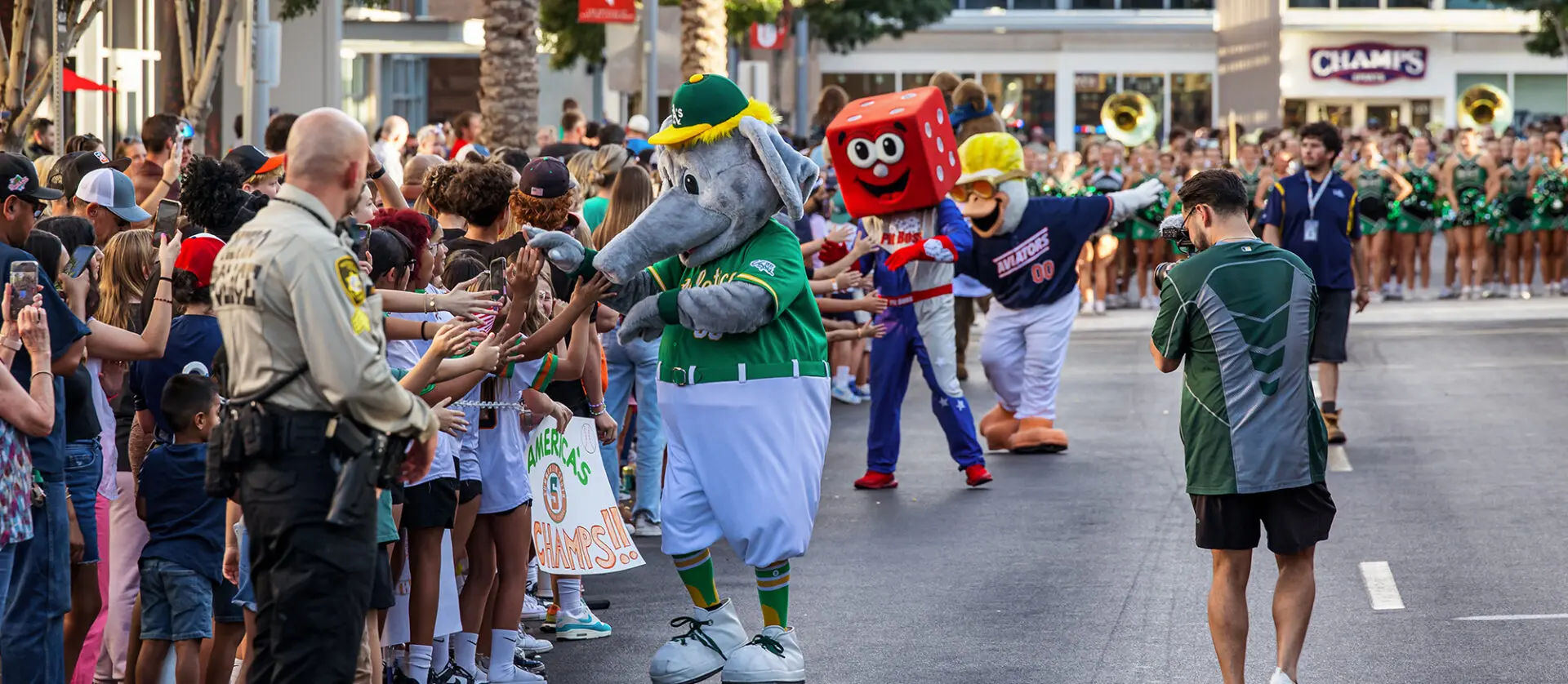 A crowd lines a city street as people in animal and dice costumes high-five spectators; a police officer and photographer are present, with a marching band and cheerleaders in the background.