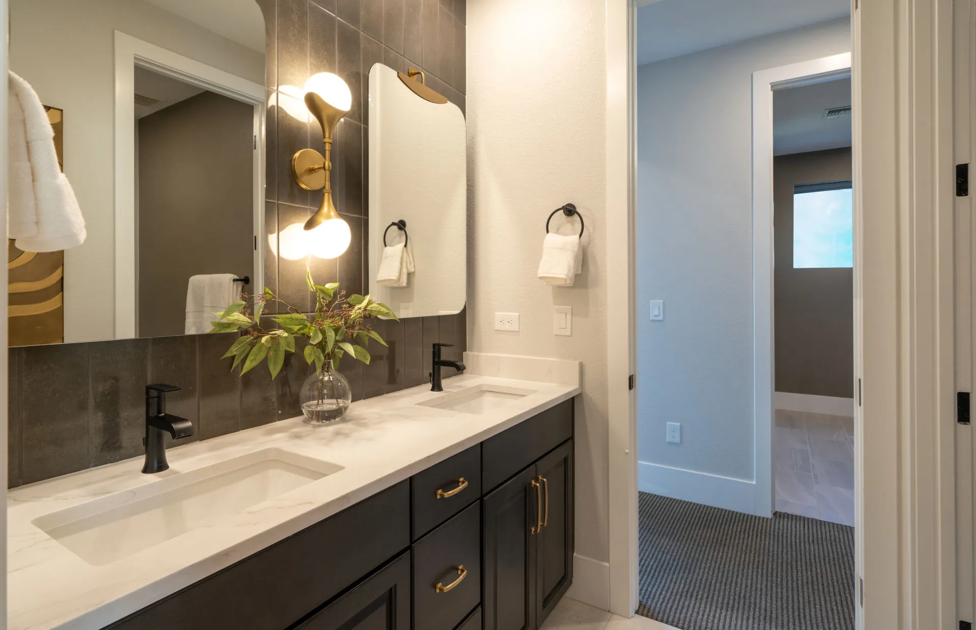 Brantley-Tivoli-Bathroom 3 Modern bathroom at Tivoli at Brantley by Pulte Homes features a double sink vanity, black cabinets, marble countertop, gold fixtures, and a glass vase with greenery. Towels hang by each sink beneath a large mirror above the vanity.