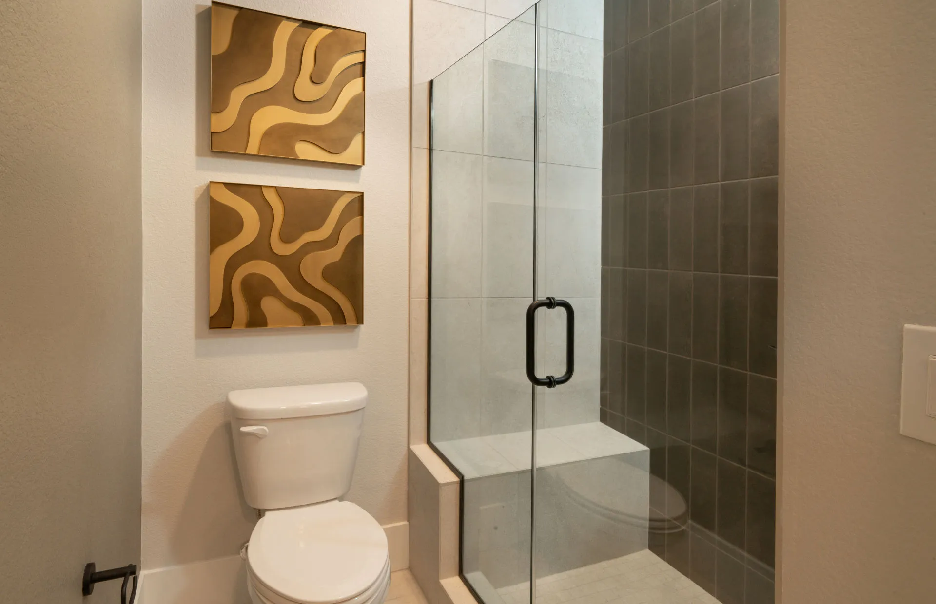 Brantley-Tivoli-Bathroom 3c A modern bathroom in Tivoli at Brantley by Pulte Homes features a white toilet, two abstract gold wall art pieces above it, and a glass-enclosed shower with dark gray tiles and a built-in bench.