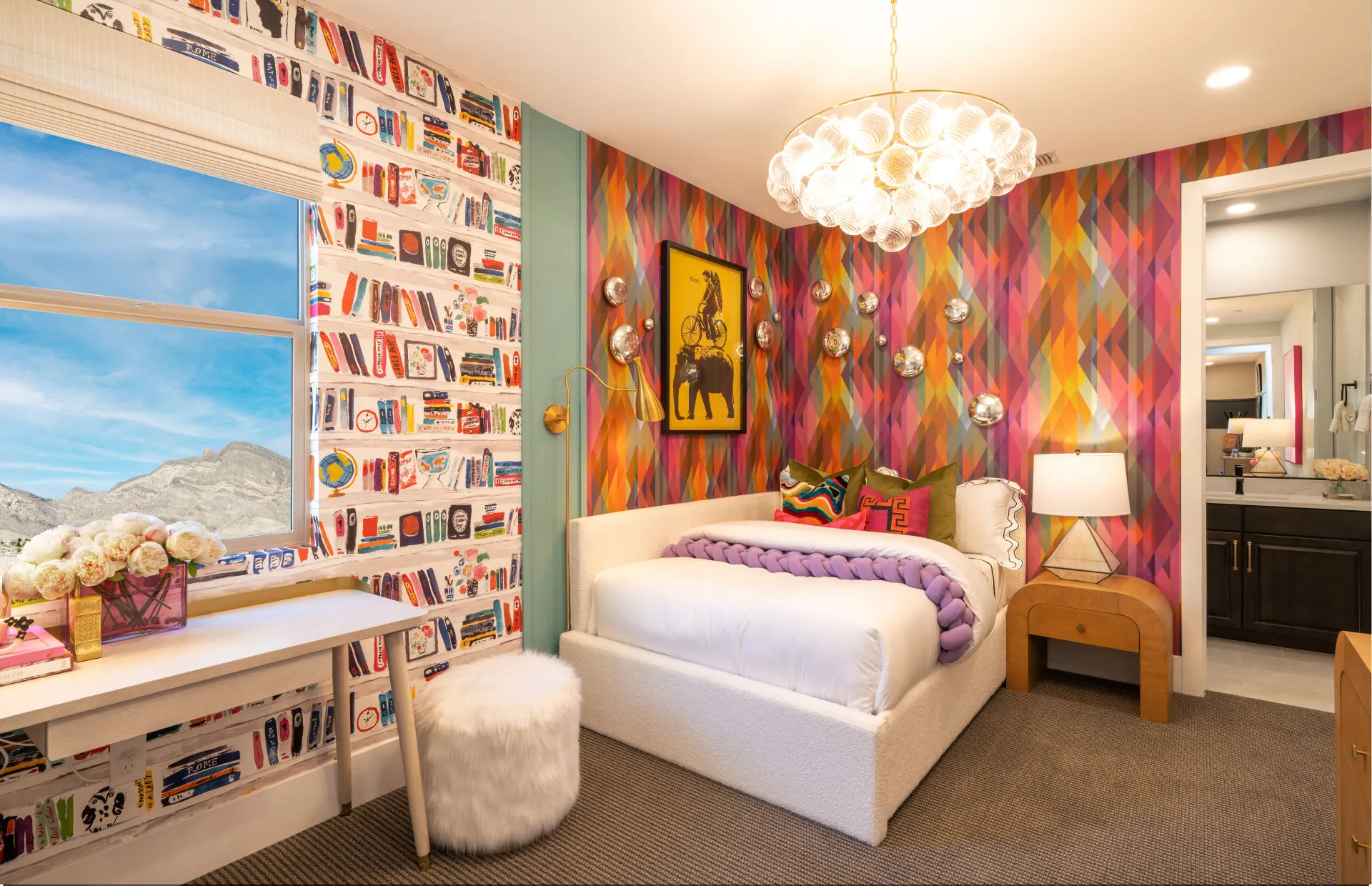 Brantley-Tivoli-Bedroom 2 A colorful bedroom at Tivoli at Brantley by Pulte Homes features a white bed with a purple knit blanket, books-themed and geometric wallpaper, a modern chandelier, desk by the window, furry white stool, and an open door to the bathroom.