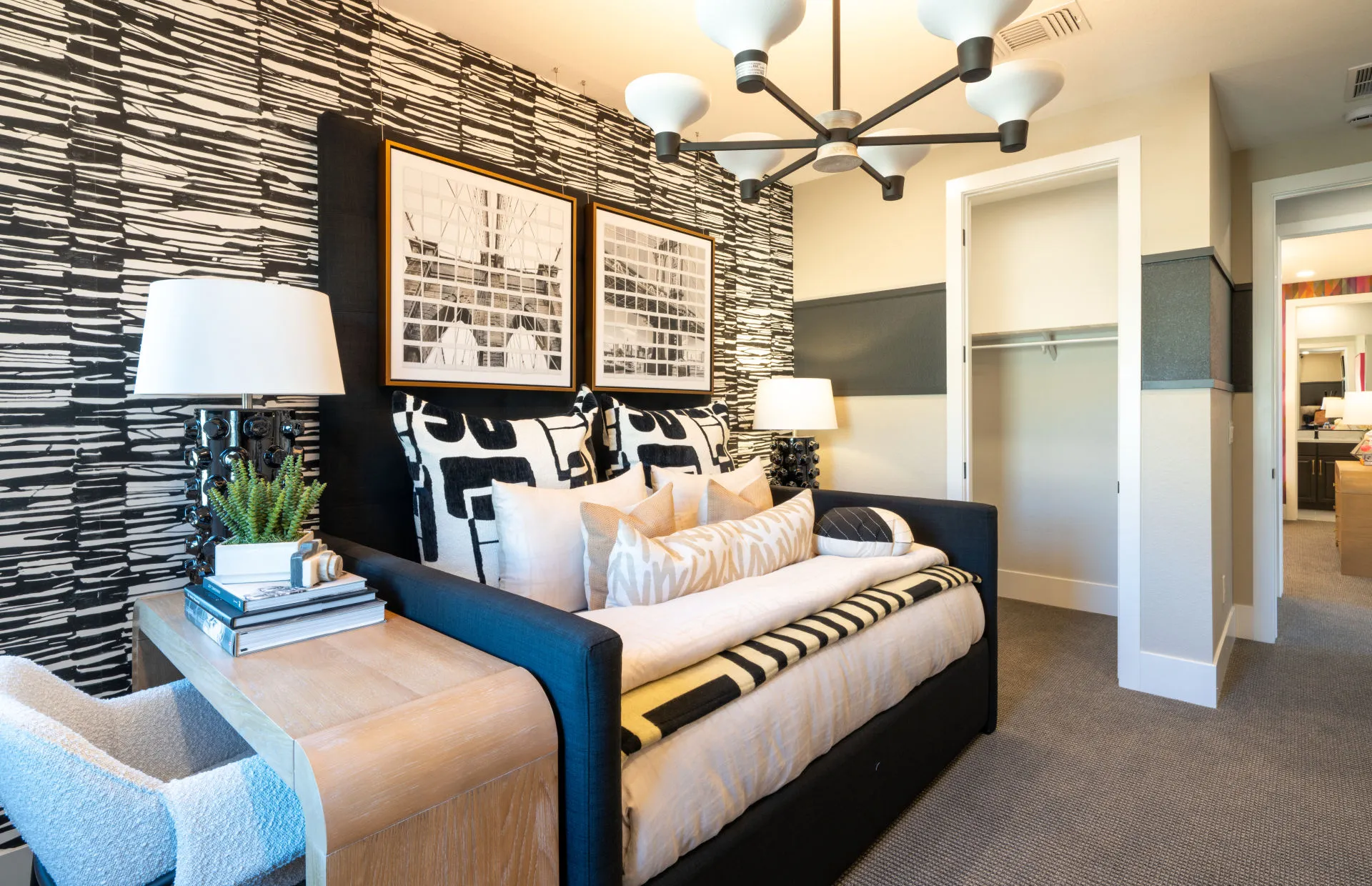 Brantley-Tivoli-Bedroom 2b Modern bedroom at Tivoli at Brantley by Pulte Homes featuring a black and white geometric accent wall, a daybed with patterned cushions, a wooden side table with books and a plant, abstract prints, and an open closet in the background.