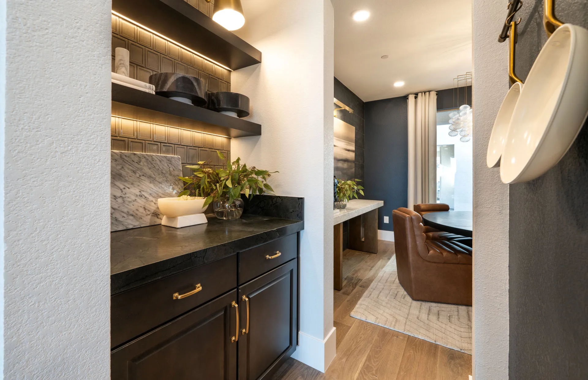 Brantley-Tivoli-Butler Pantry A modern home interior at Tivoli at Brantley by Pulte Homes features black cabinets with gold handles, open shelves with decor, and indoor plants. In the background, a cozy seating area boasts a brown leather chair and a large rug on wood flooring.