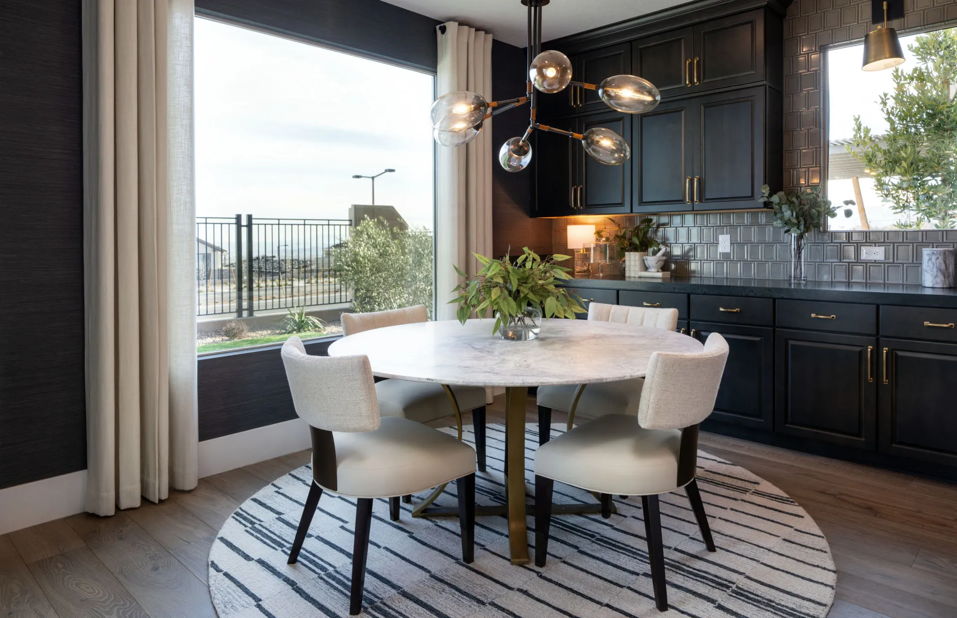 Brantley-Tivoli-Cafe A modern dining area at Tivoli at Brantley by Pulte Homes features a round marble table, four upholstered chairs, a striped circular rug, black cabinets, and a contemporary chandelier. Large windows and a sliding door bring in natural light.