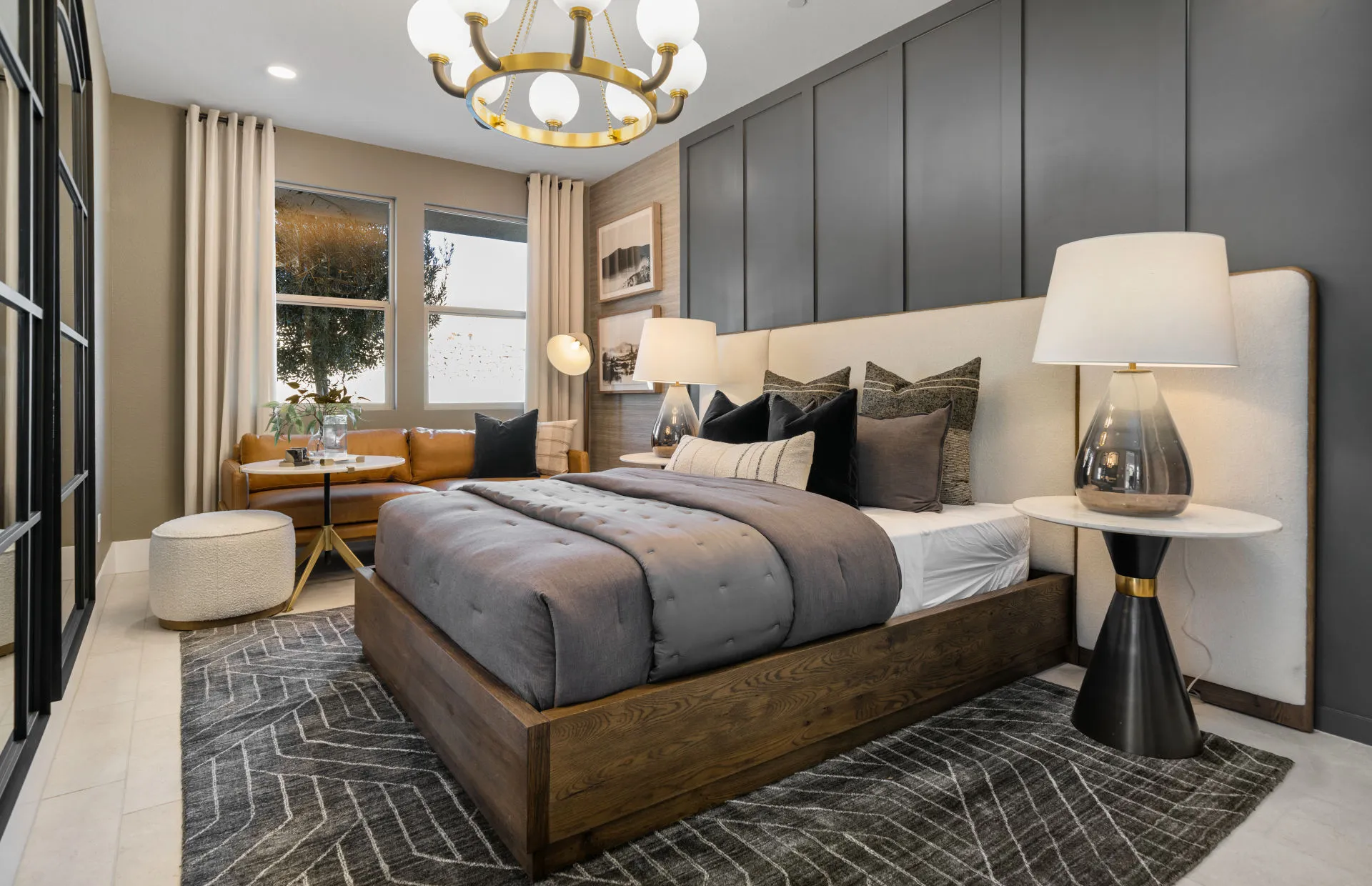 Brantley-Tivoli-Casita 1 Modern bedroom at Tivoli at Brantley by Pulte Homes features a wooden bed, gray bedding, large headboard, geometric rug, and elegant lamps. A brown sofa sits by the window with a round ottoman and a contemporary gold chandelier overhead.