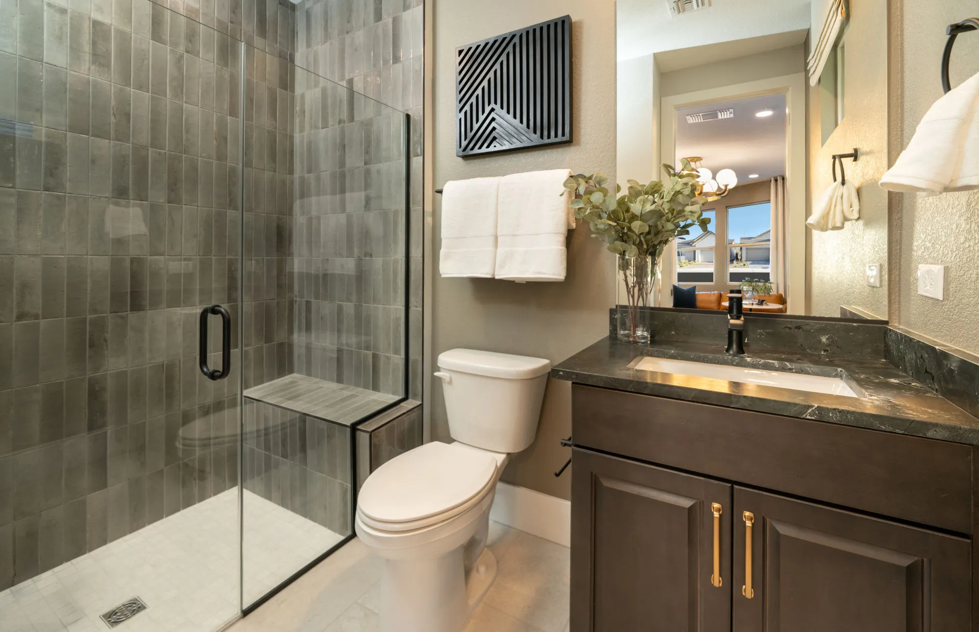 Brantley-Tivoli-Casita Bath Modern bathroom at Tivoli at Brantley by Pulte Homes features a glass-enclosed shower, white toilet, dark wood vanity with black countertop, rectangular sink, wall mirror, towel rack, and greenery. Dining area visible through doorway.