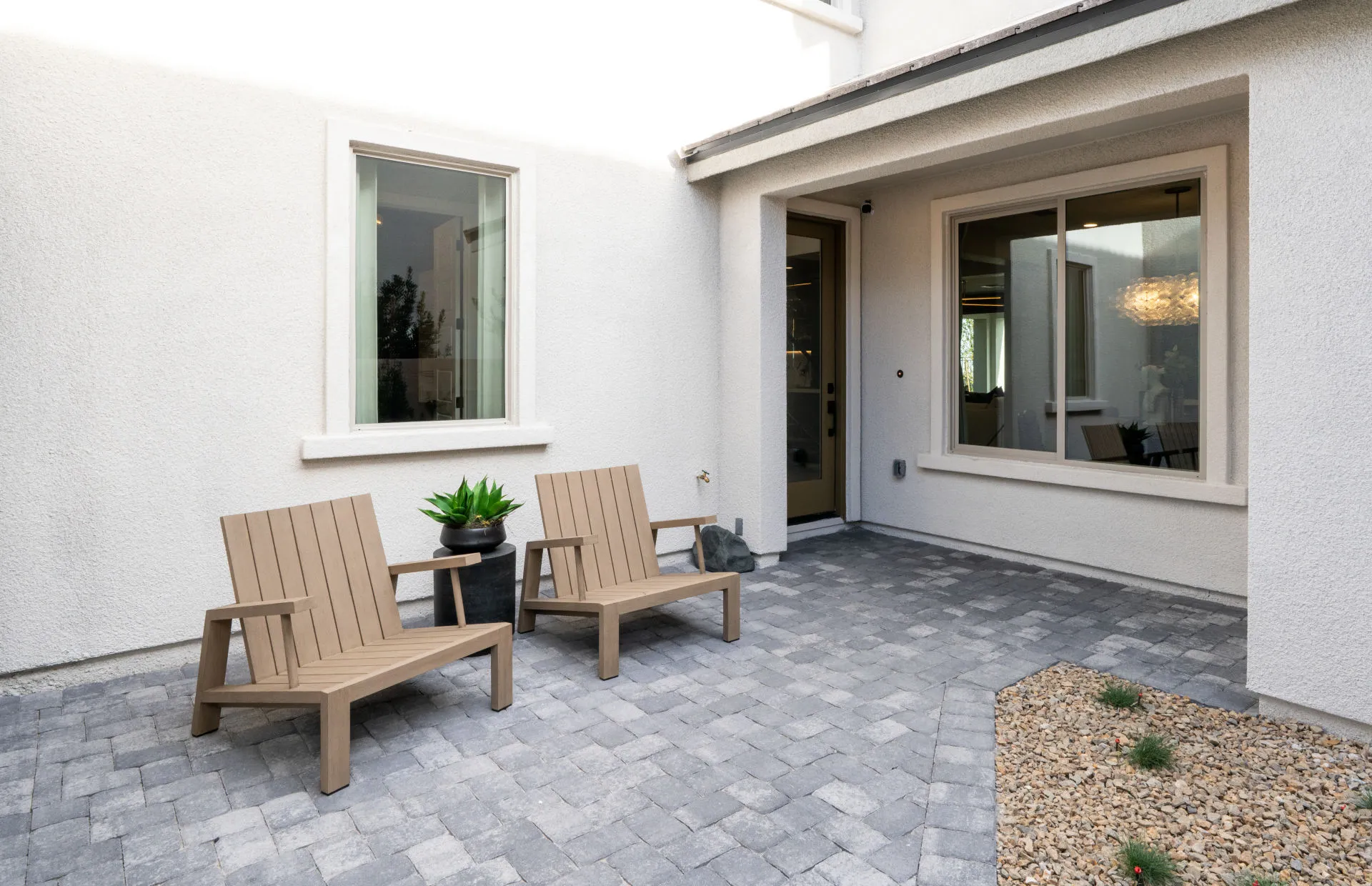 Brantley-Tivoli-Courtyard A modern patio at Tivoli at Brantley by Pulte Homes features two wooden chairs, a black side table with a potted plant, gray paver flooring, a gravel area with small plants, and white exterior walls with large windows.