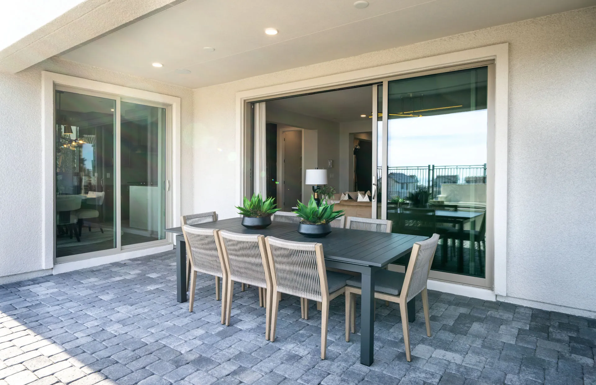 Brantley-Tivoli-Covered Loggia A modern outdoor patio at Tivoli at Brantley by Pulte Homes features gray pavers, a rectangular dining table with six chairs, and two potted plants. Sliding glass doors open to a stylish interior with views of the fenced backyard.