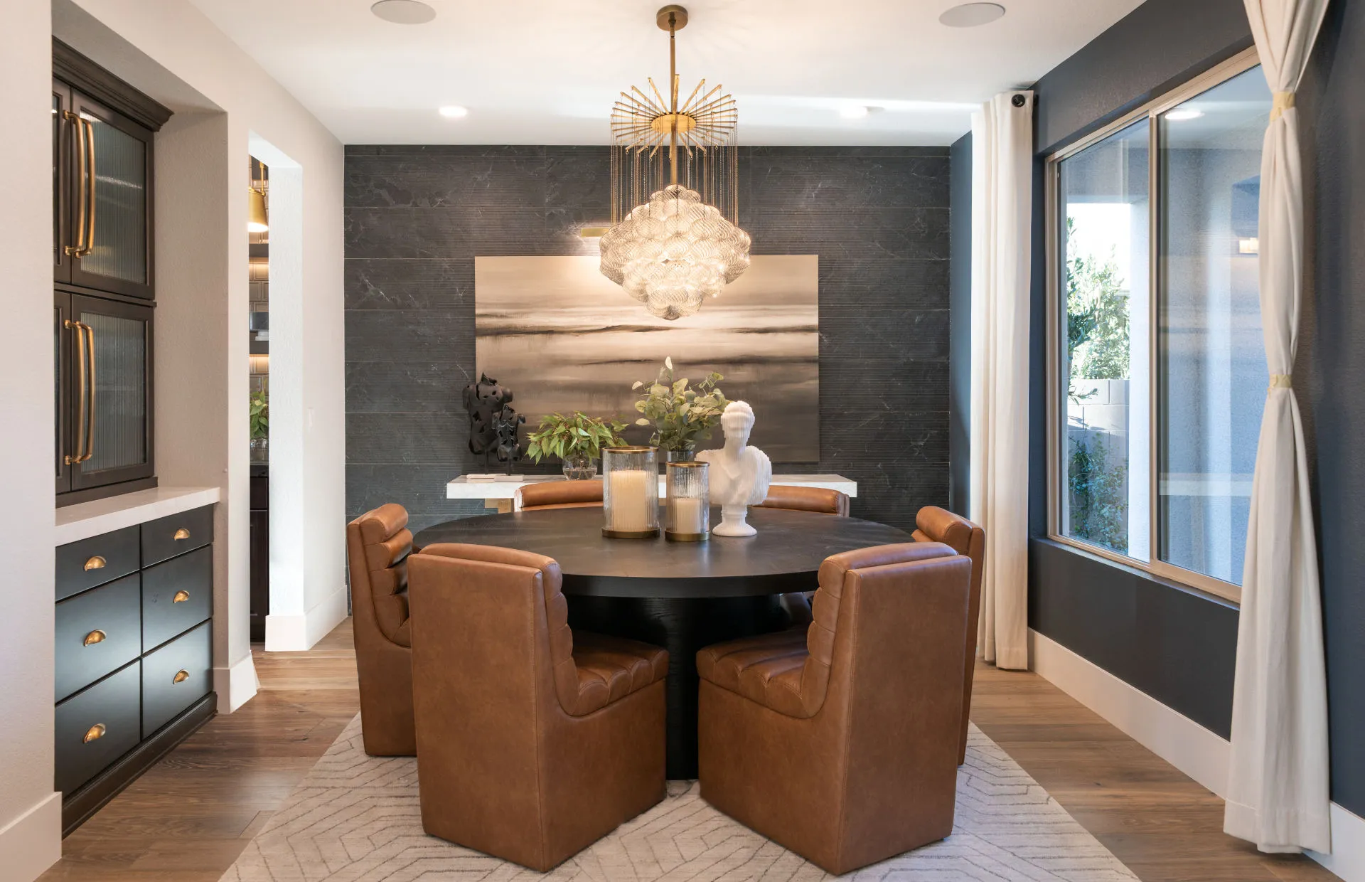Brantley-Tivoli-Dining A modern dining room at Tivoli at Brantley by Pulte Homes features a round black table, six brown leather chairs, a contemporary chandelier, abstract wall art, decorative plants, and large windows with sheer curtains letting in natural light.