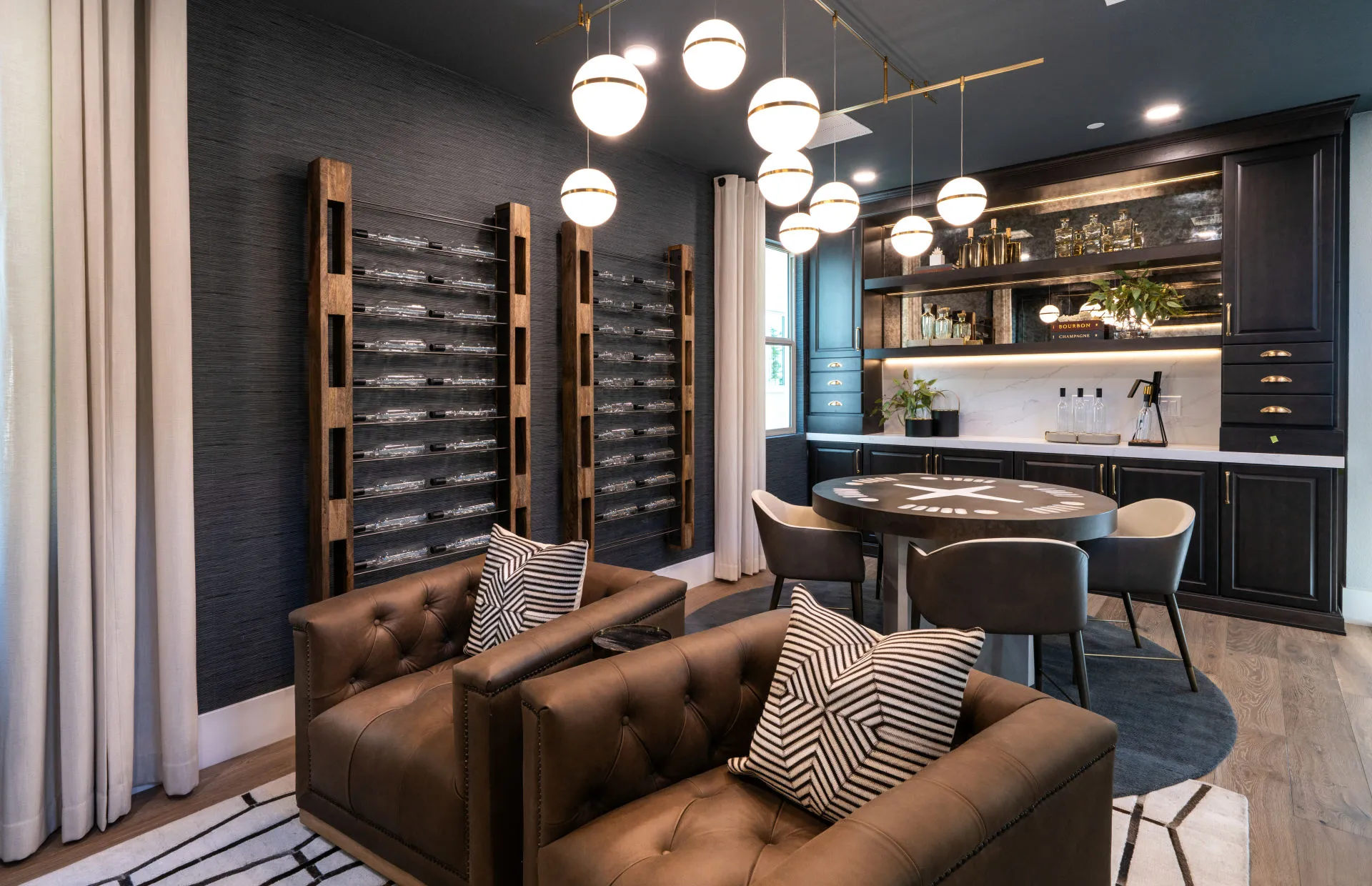 Brantley-Tivoli-Flex Room 2 Modern lounge at Tivoli at Brantley by Pulte Homes features tufted leather chairs, geometric pillows, a round poker table, contemporary chandelier, wall-mounted wine racks, and a sleek bar area with dark cabinetry and stylish decor.