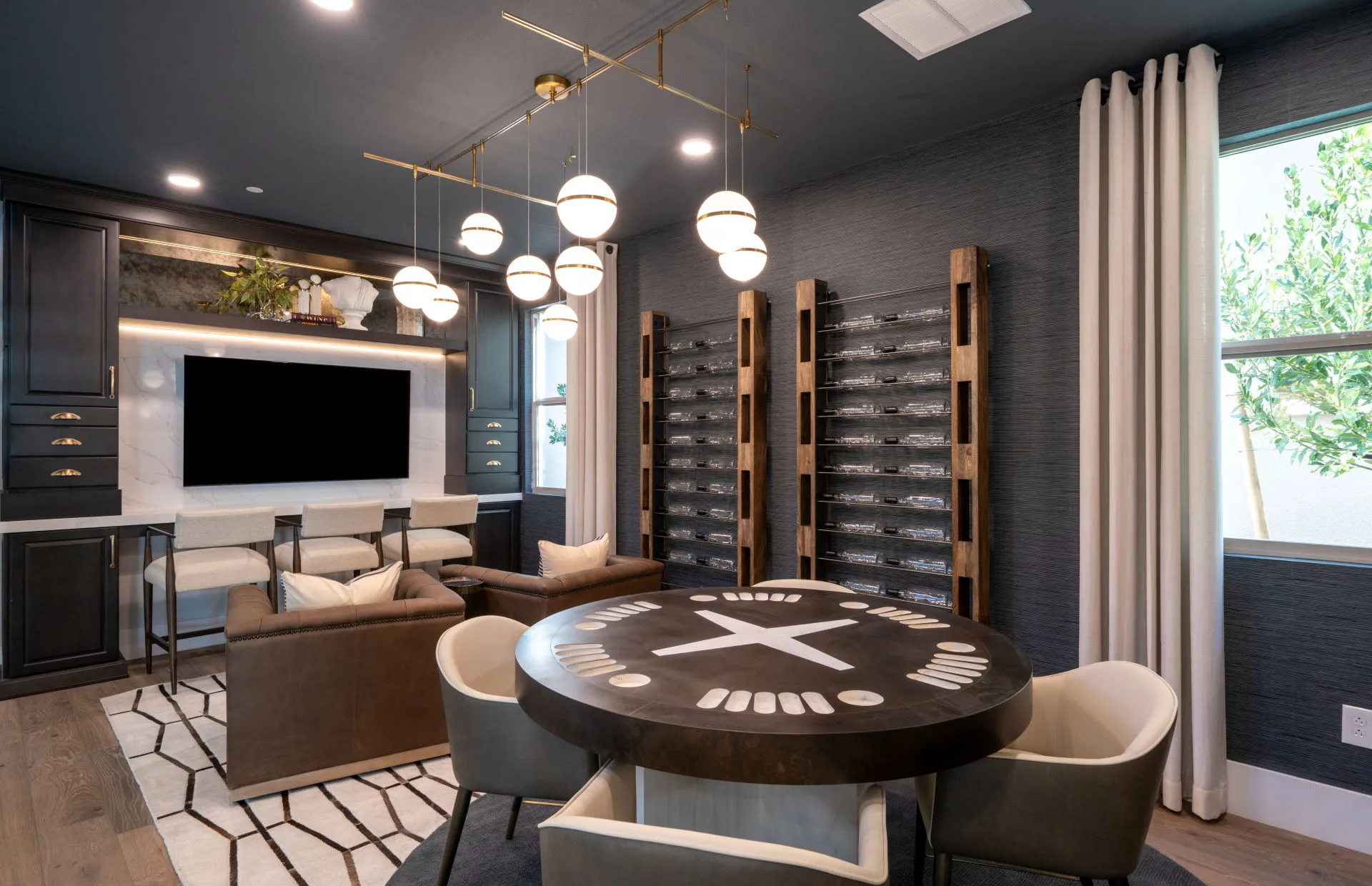 Brantley-Tivoli-Flex Room Modern entertainment room at Tivoli at Brantley by Pulte Homes features a round game table, plush chairs, built-in TV, sofa, and shelving. Pendant lights hang from the dark ceiling, with large windows and white curtains welcoming natural light.