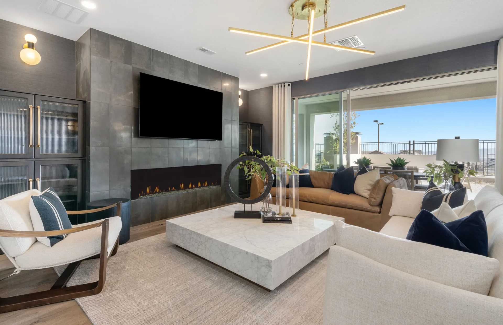 Brantley-Tivoli-Gathering Room 2 Modern living room at Tivoli at Brantley by Pulte Homes features a marble coffee table, beige and white sofas, mounted TV above a sleek fireplace, large windows with outdoor views, gold chandelier, and contemporary decor.