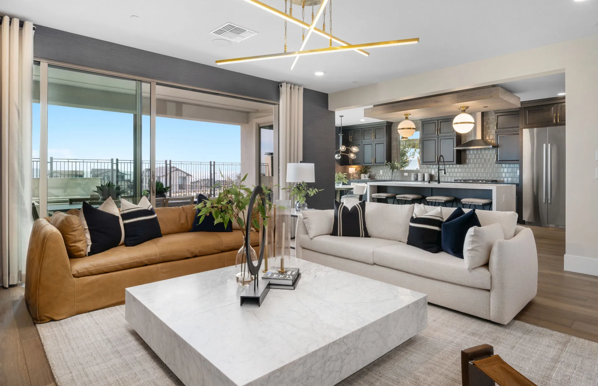 Brantley-Tivoli-Gathering Room1 Modern living room at Tivoli at Brantley by Pulte Homes, featuring tan and cream sofas, a marble coffee table, decorative pillows, and views into a stylish kitchen. Glass doors open to a patio with outdoor seating.