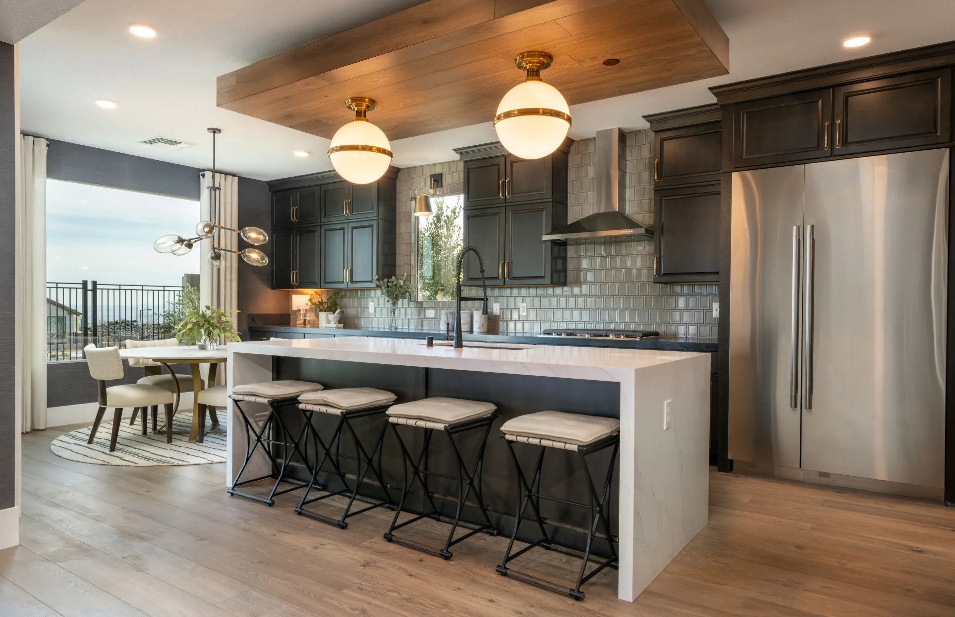 Brantley-Tivoli-Kitchen Cafe Modern kitchen at Tivoli at Brantley by Pulte Homes features dark cabinets, a stainless steel refrigerator, and a white island with four barstools under pendant lights. A dining area with a round table sits near large windows.