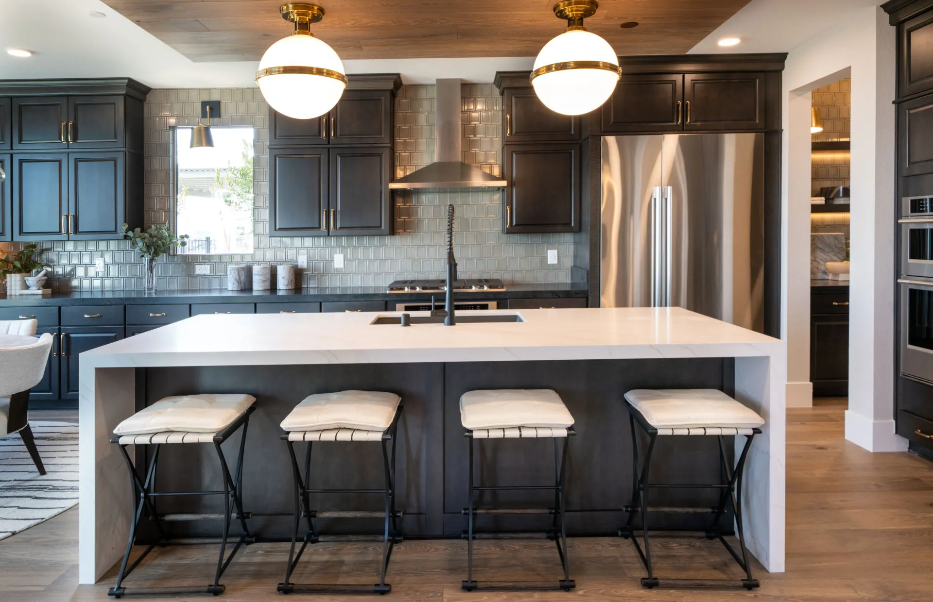 Brantley-Tivoli-Kitchen Modern kitchen with dark cabinets, stainless steel appliances, a large white island with four cushioned stools, globe pendant lights, wood floors, and a neutral color palette at Tivoli at Brantley by Pulte Homes.