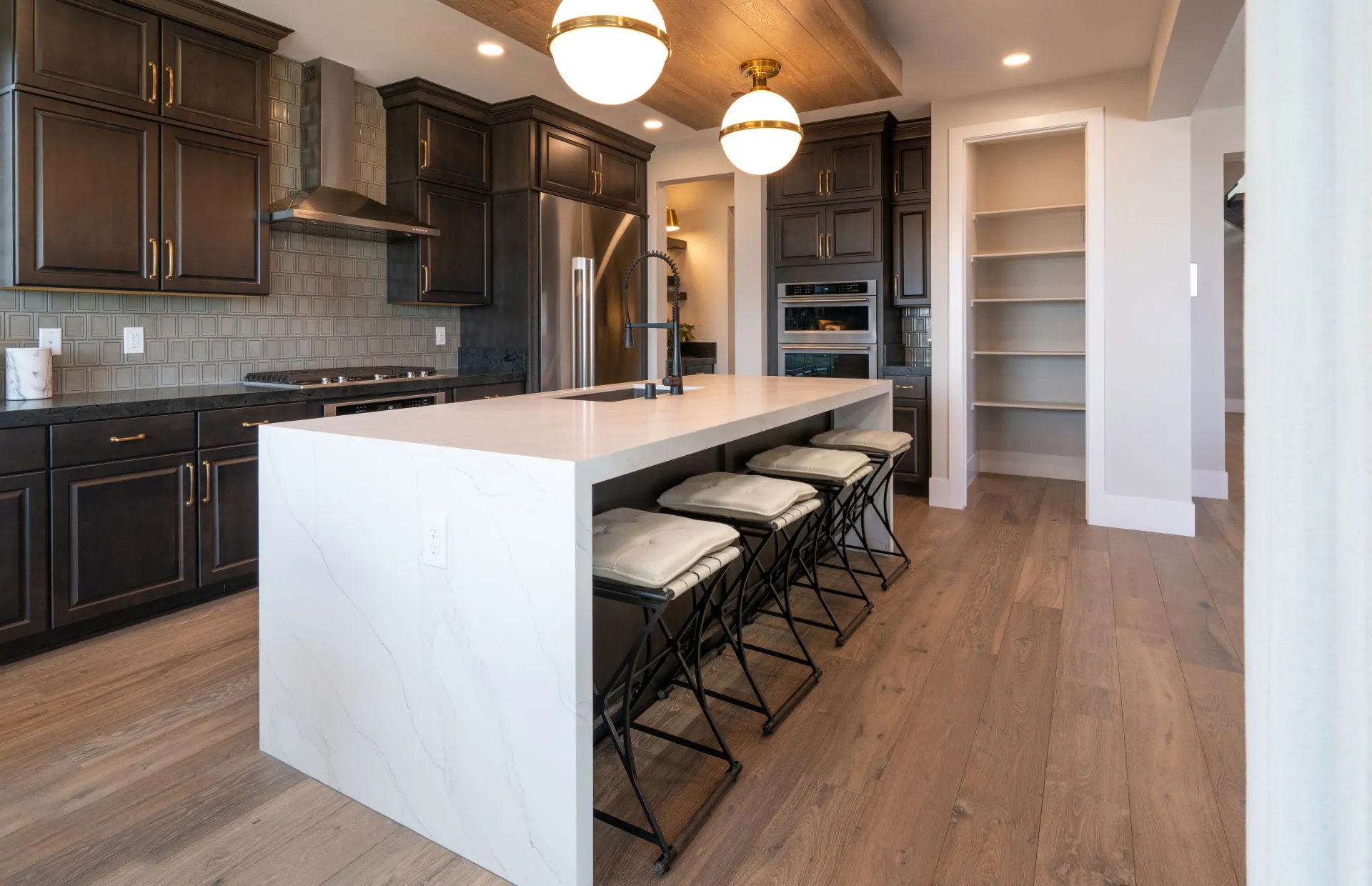 Brantley-Tivoli-Kitchen2 Modern kitchen at Tivoli at Brantley by Pulte Homes featuring dark cabinets, stainless steel appliances, light wood flooring, a large white marble island with built-in sink, four cushioned stools, open shelves, and pendant lights.